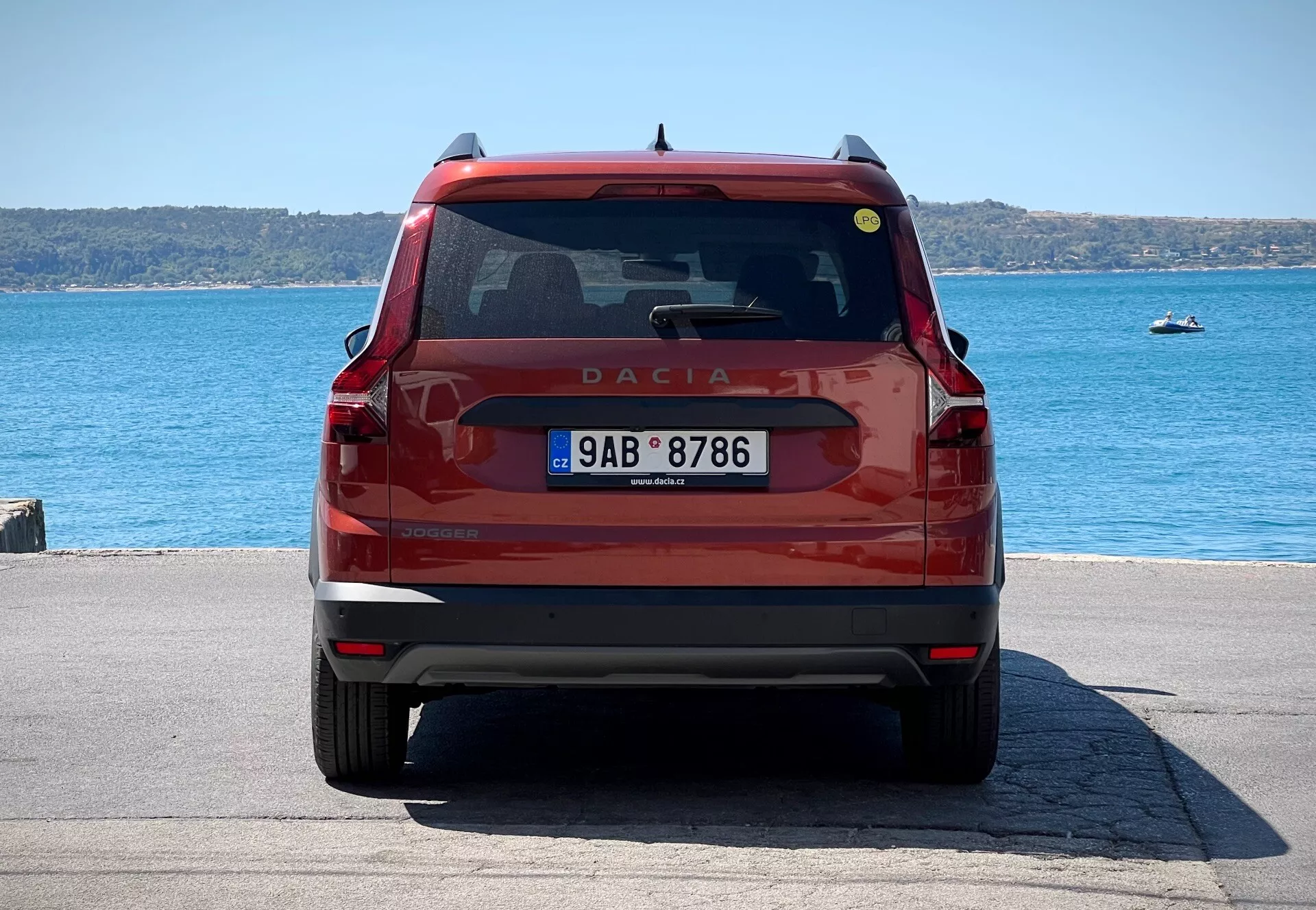 Zadní pohled na červený Dacia Jogger 2022, předvádějící zadní dveře s emblémem a názvem modelu, zadní čelní sklo s stěračem a brzdovým světlem, moderní zadní světla, registrační značku a nárazník s odrazkami a možnými parkovacími senzory, zaparkovaný u nábřeží ve dne.