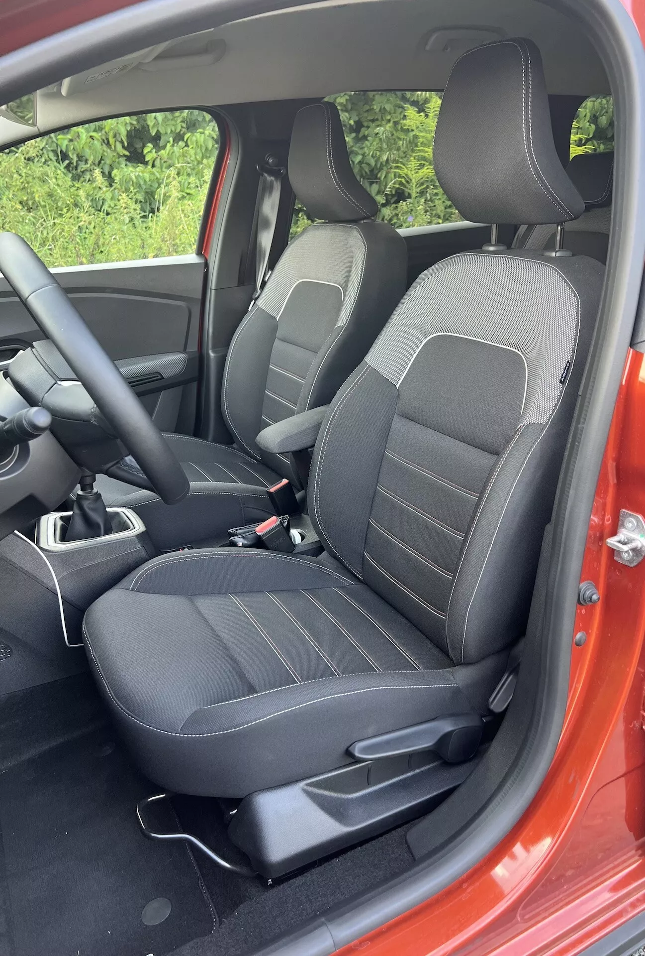 Podrobný interiérový pohled na Dacia Jogger 2022 ukazující přední prostor pro cestujícího s černě čalouněnými sedadly s lineárními vzory, ovládacími prvky pro nastavení, částí středové konzoly a náznakem červeného exteriéru