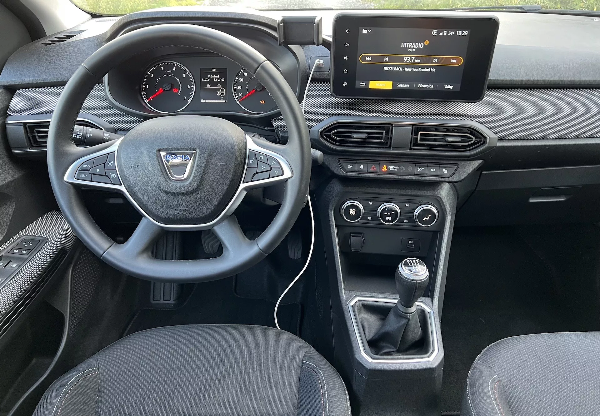 Interiérový pohled na Dacia Jogger 2022 ze zadního sedadla, zvýrazňuje palubní desku řidiče, volant s logem výrobce a ovládacími tlačítky, digitálně-analogový přístrojový panel, dotykový informační systém, ovládání HVAC, klimatizaci s duálním nastavením a manuální převodovku