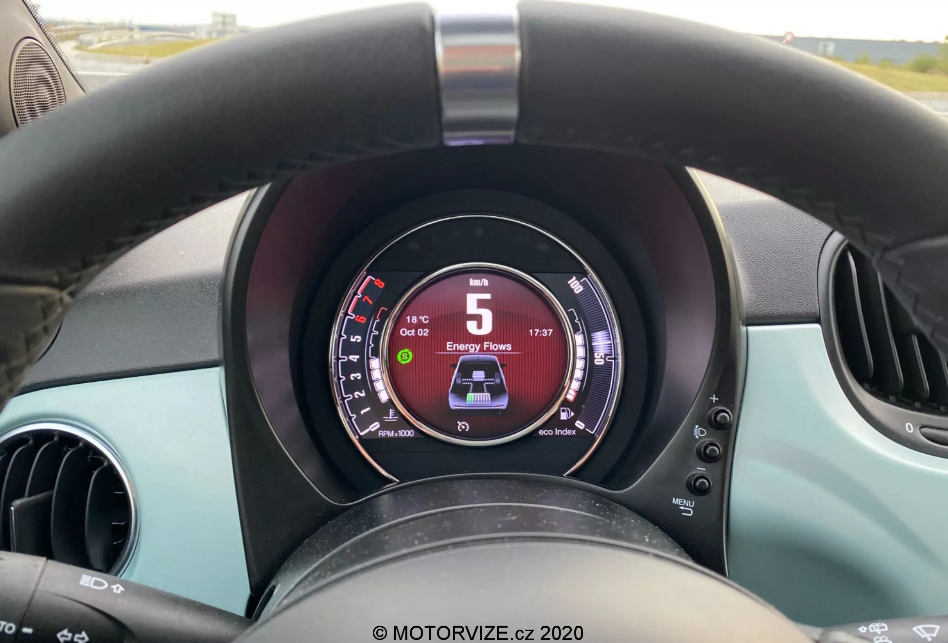 Detailní pohled z pohledu řidiče na interiér Fiat 500 2015, zvýrazňující černý lehce opotřebovaný volant, digitální panel přístrojů ukazující rychlost 5 km/h a 'D' pro jízdu vpřed, dva kruhové ventilační otvory a tyrkysové akcenty na palubní desce a dveřích.