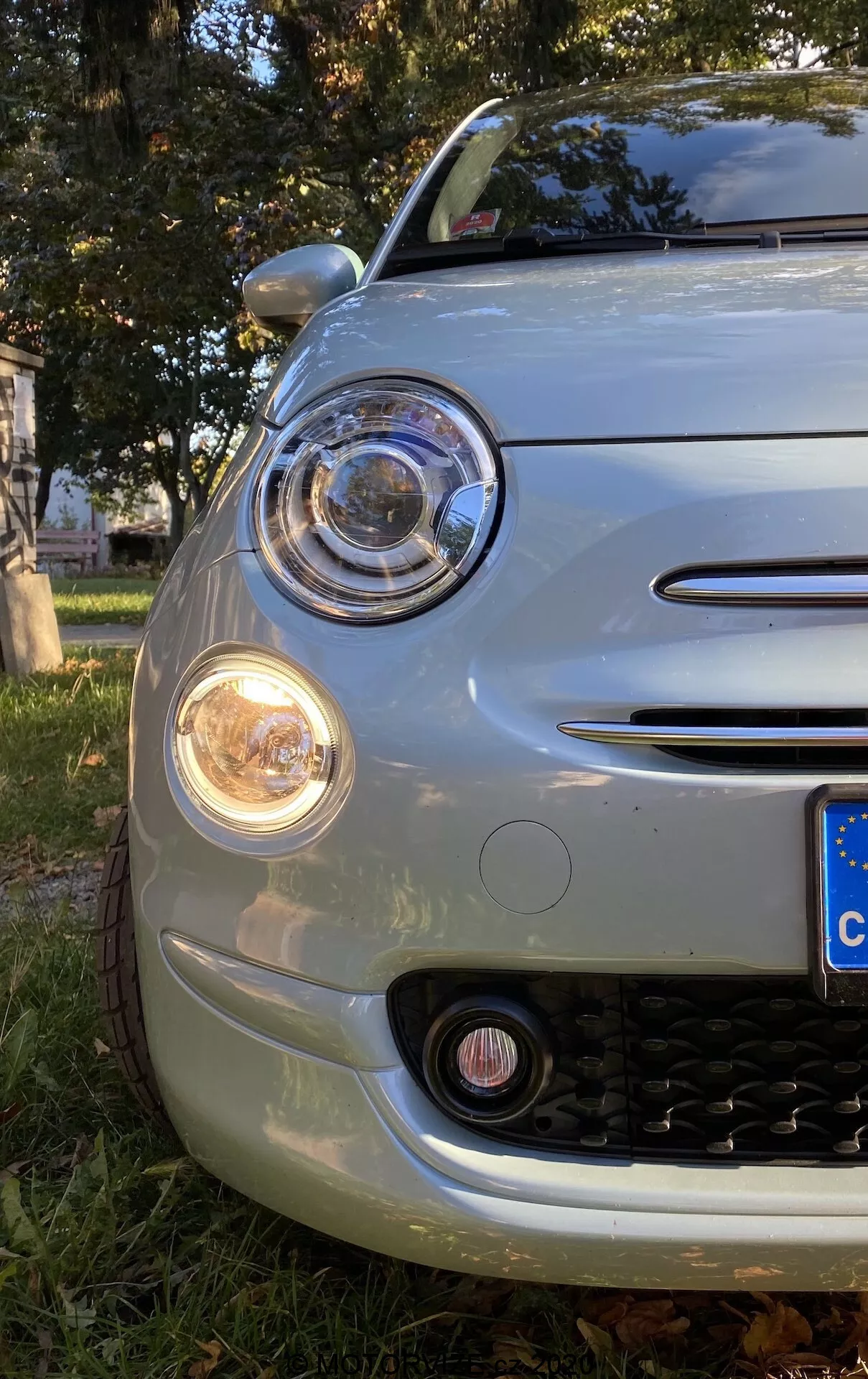 Detailní pohled ze spodního úhlu na přední levý roh modelu Fiat 500 2015, zobrazující kapotu, přední nárazník, oblouk kola a kruhový hlavní světlomet s projektorem obklopeným denními světly (DRL). Světelný komplet obsahuje signálku v oranžové barvě a mlhové světlo dole. Chromované akcenty zdobí okraj světlometu, kryt mlhových světel a dva horizontální proužky na mřížce. Mřížka má černý voštinový vzor. Kulatá přední část a evropská SPZ ukazují retro-moderní design auta, zaparkovaného na trávě s podzimními listy.