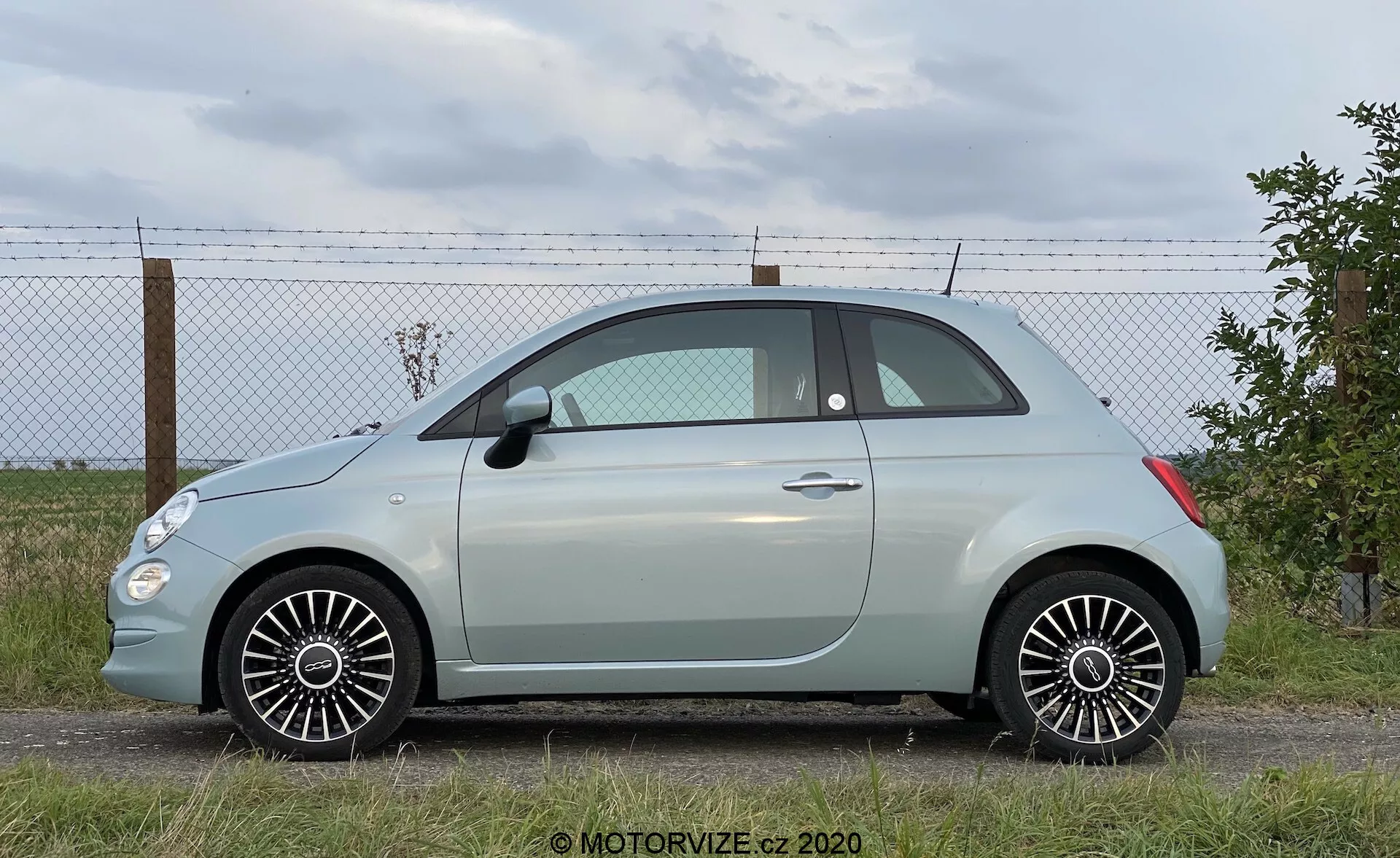 Moderní subkompaktní hatchback z bočního úhlu, zobrazující celou délku vozidla. Zaparkováno na rovném povrchu s obvodovým plotem a horizontem s oblaky v pozadí. Obsahuje boční dveře, okna, zrcátka, dvoudveřovou konfiguraci, malé zadní čtvrtokno, zaoblenou střechu, pastelově zbarvenou karoserii s kontrastní střechou, vícepaprskové slitiny ráfků, pneumatiky v dobrém stavu, detailní designové linie a přirozené denní osvětlení.