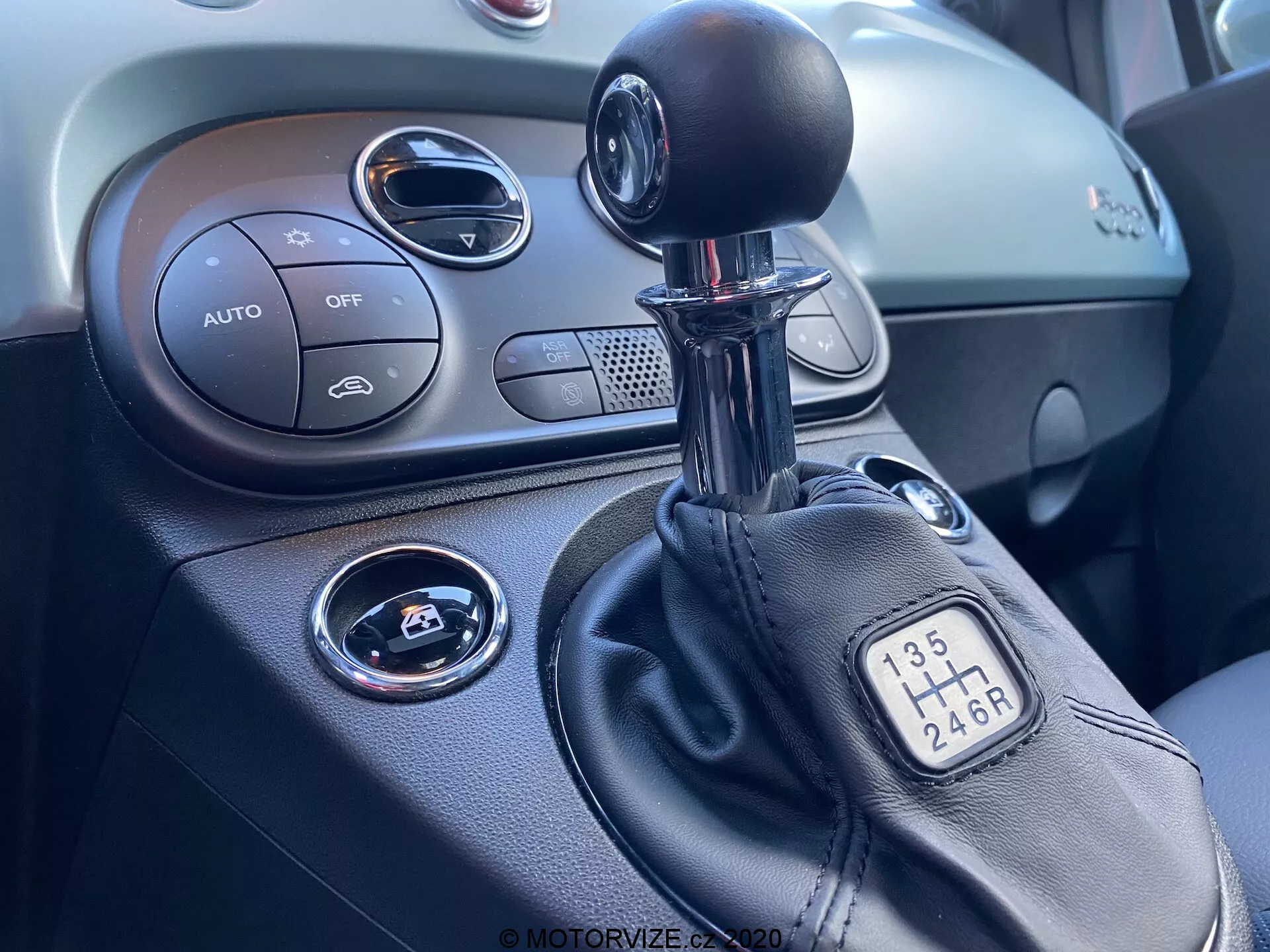 Detailní pohled na středovou konzoli uvnitř vozu Fiat 500 z roku 2015, zaměřený na pravou stranu. Obraz ukazuje manuální řadicí páku s konfigurací 6 rychlostí, část středové konzole s ovládáním klimatizace označeným 'AUTO', tlačítka 'OFF', tlačítkem start/stop pro zapalování, kruhový vzduchový ventil a texturovanou palubní desku.