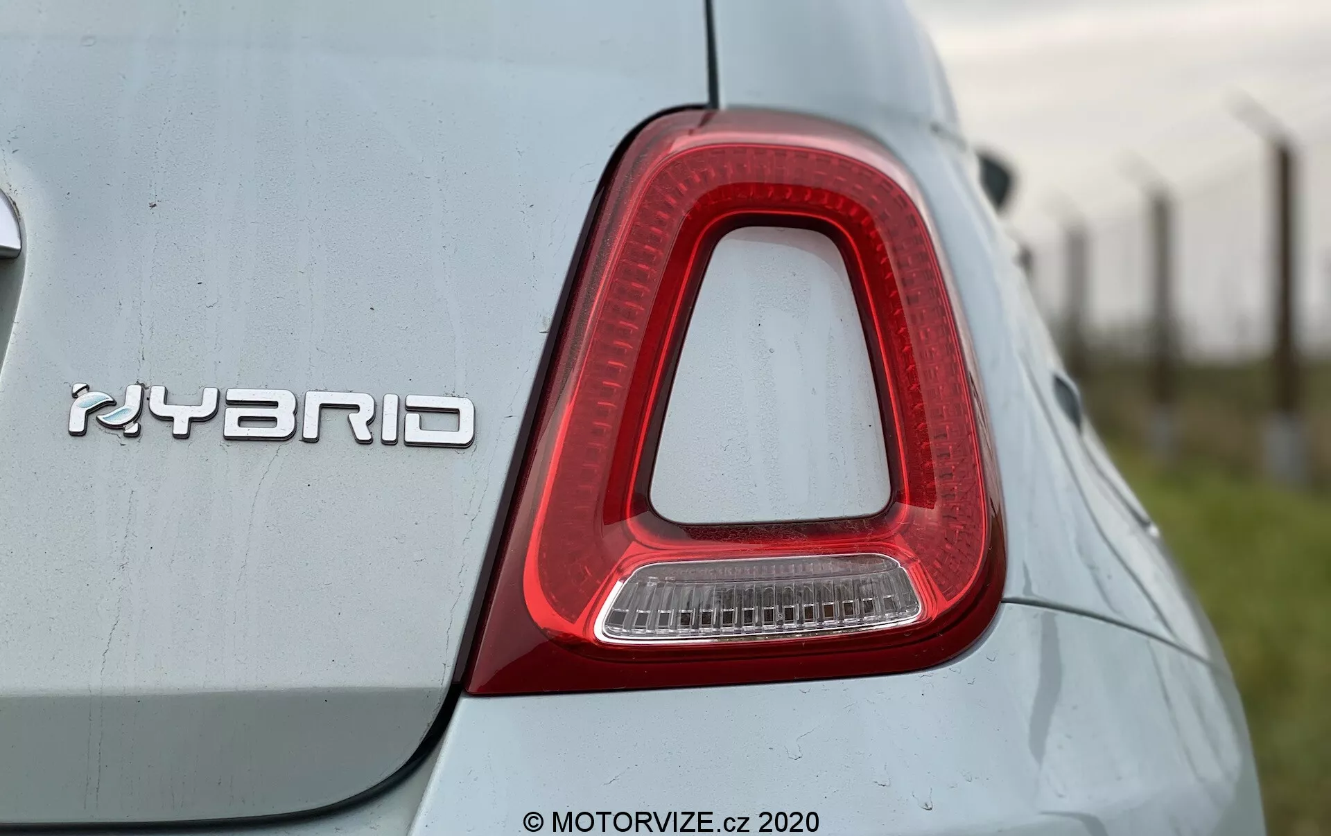 Detailní záběr na pravé zadní světlo a odznak 'HYBRID' na Fiat 500 2015. Zadní světlo má tvar obráceného L, převážně červené s průhlednou částí pro couvací světlo a je zarovnané s karoserií vozu. Kovový odznak 'HYBRID', indikující hybridní pohon, je výrazně umístěn. Světlá barva karoserie má odrazivou kvalitu s prachovými nebo vodními skvrnami, což naznačuje venkovní prostředí. Obrázek je pořízen z mírně nízkého úhlu, zvýrazňující design zadního světla a hybridní technologii.
