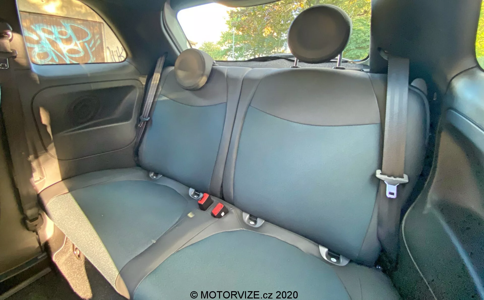 Interiér vozu Fiat 500 z roku 2015 zobrazující zadní sedadla s čalouněním z kůže nebo koženky v tmavé barvě, viditelné švy, tříbodové bezpečnostní pásy, nastavitelná opěrky hlavy, dveřní panel s ovládáním elektrického okna, úložný prostor, mřížku reproduktoru, částečný pohled na strop a horní držadlo. Úhel kamery je mírně zvýšený zepředu dozadu, poskytující podrobný pohled na zadní lavicové sedadlo, zadní dveře, část stropu a okna.