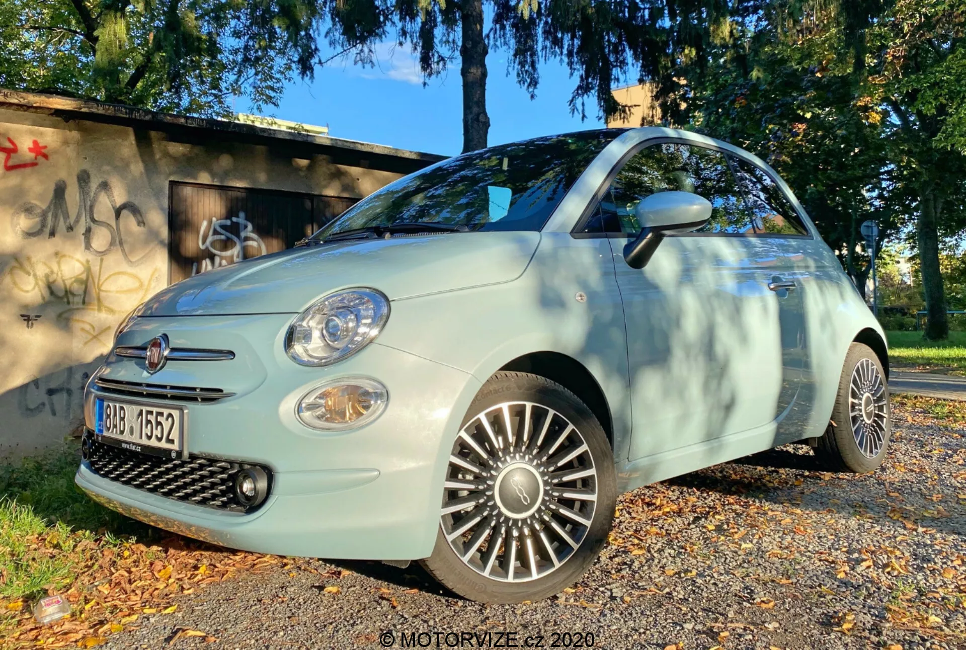 Záběr z nízkého úhlu na Fiat 500 2015 z předního čtvrtletí, ukazující řidičovu stranu. Obraz zvýrazňuje mřížku, přední nárazník, světlomety, mlhovky a podrobné slitá kola. Auto je zaparkováno na povrchu pokrytém podzimními listy, s travnatou plochou a strukturou pokrytou graffiti v pozadí, zalité přirozeným denním světlem, což naznačuje ranní nebo pozdní odpolední osvětlení.