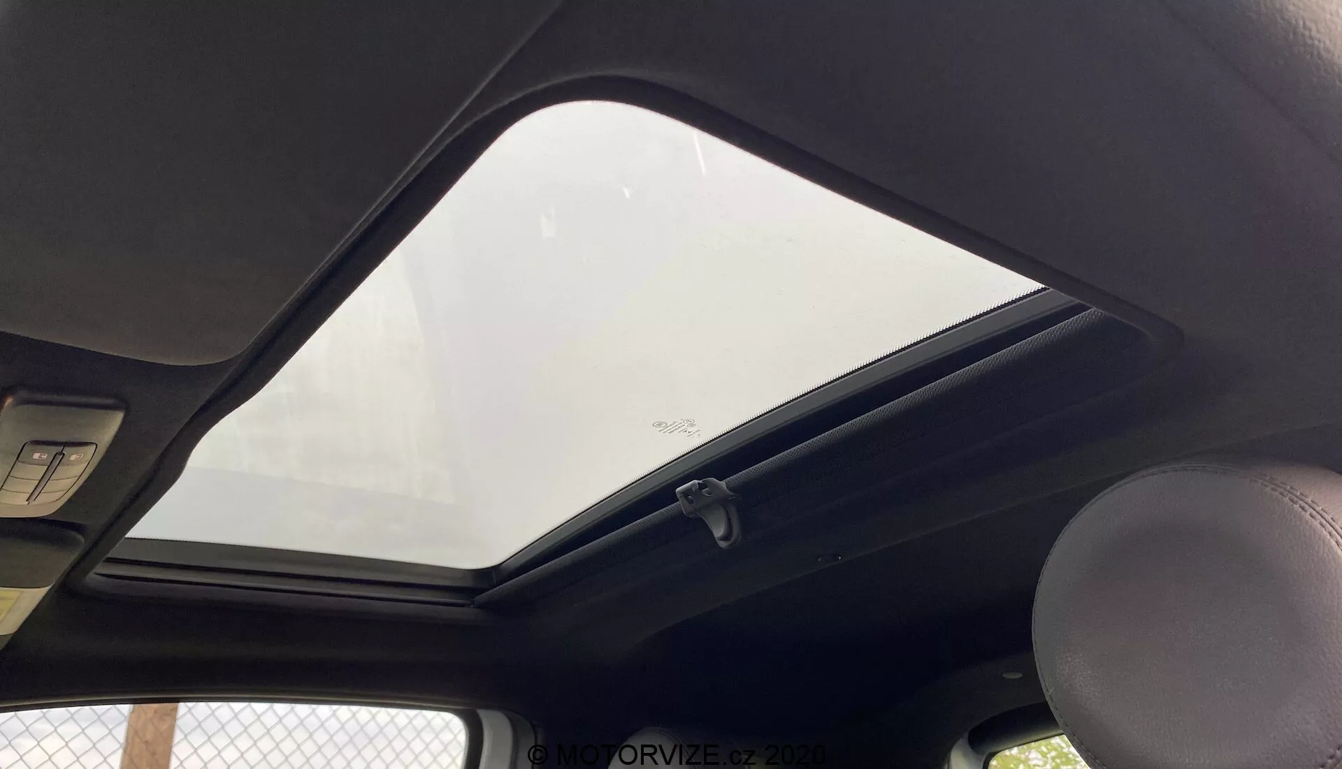Interiér Fiat 500 2015 s pohledem zaměřeným na horní část směrem k střeše z perspektivy pasažéra nebo řidiče. Výrazný uzavřený střešní okno umožňuje průnik přirozeného světla do kabiny. Tmavá stropní látka kontrastuje s exteriérem. Viditelná madlo na levé straně, standardní stropní světlo nad ním a okraje dvou slunečních clon nahoře. Háček na oblečení je u hranice střešního okna, které má na spodní straně bezpečnostní štítek. Obrázek je pořízený z nízkého úhlu a blízce rámuje interiérové prvky střechy.