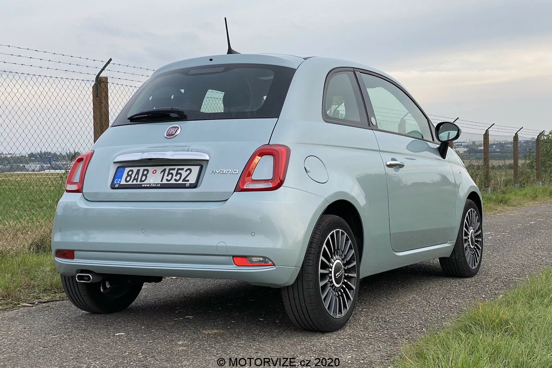 Zadní pohled z tříčtvrtinového úhlu na světle modré kompaktní auto Fiat 500 z roku 2015, který ukazuje zaoblená čtvercová zadní světla, zadní stěrač, chromovou značku, parkovací senzory na nárazníku, výfukovou trubku na levé straně a litá kola. Auto je zaparkované s levou stranou směřující k fotoaparátu, před rozmazaným plotem a zelení v pozadí.