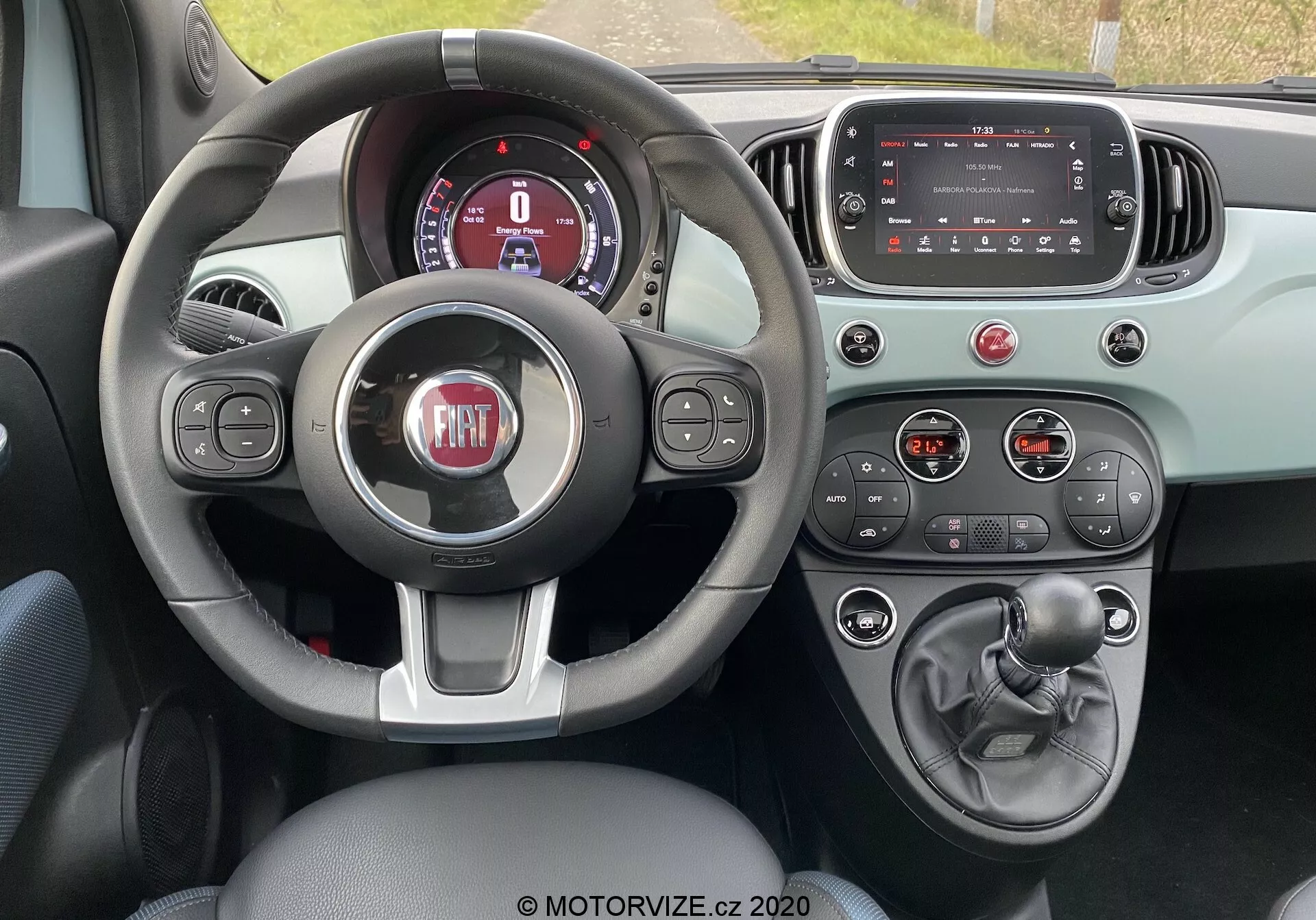 Interiér modelu Fiat 500 2015 z pohledu řidiče zobrazující tříramenný volant s ovládacími tlačítky, částečně viditelný digitální přístrojový panel, vypnutou dotykovou obrazovku infotainmentu, tlačítka a knoflíky klimatizace, manuální řadicí páku, ruční brzdu a barevnou dvoutónovou kombinaci zdůrazňující funkčnost a moderní designové prvky.