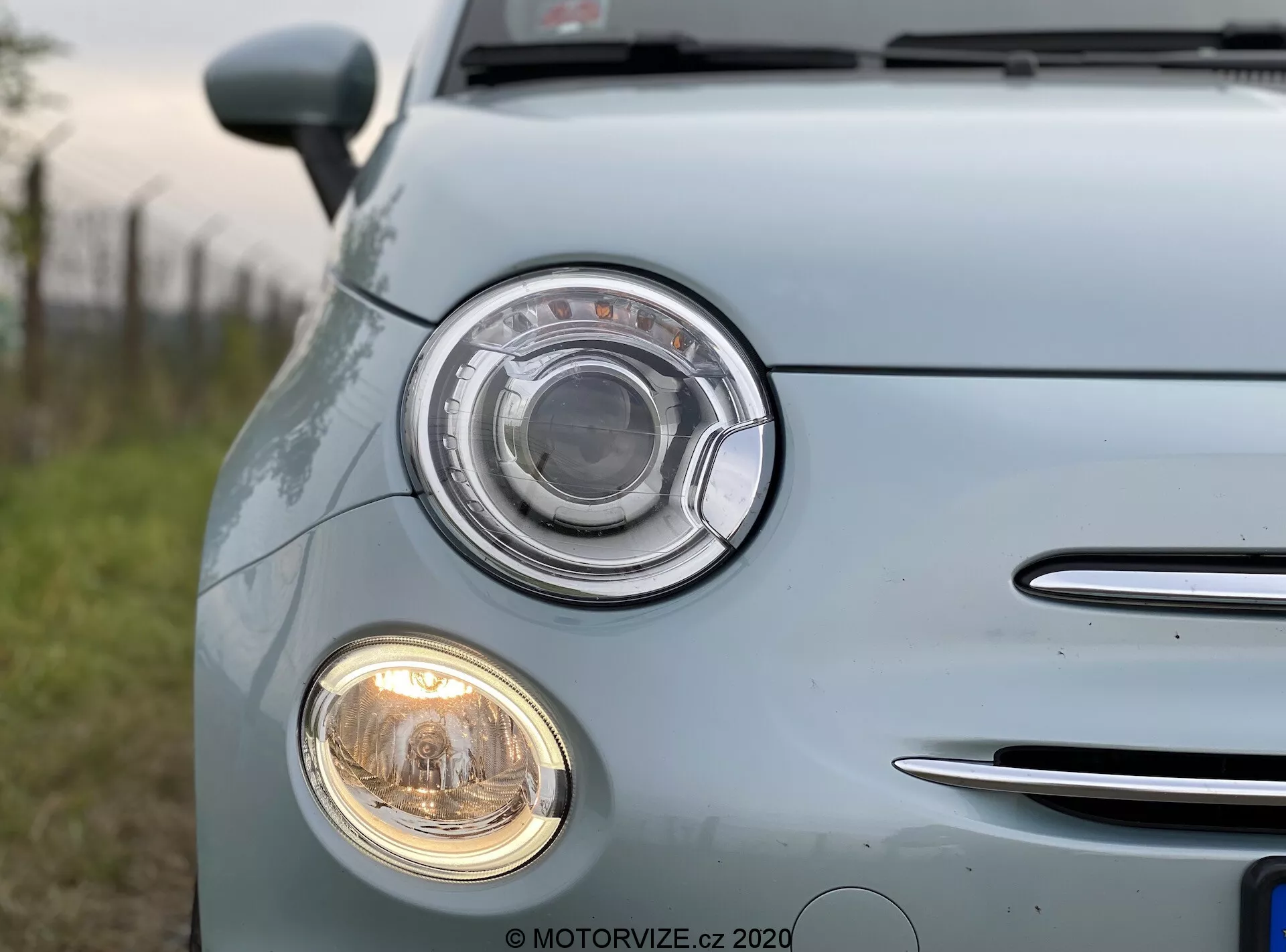 Detailní snímek světle modrého Fiatu 500 z roku 2015, pořízený ze čtvrtiny přední levé části. Zaměřeno na výrazný světlomet s kruhovým designem a čirým krytem, stejně jako menší kruhový mlhový světlomet pod ním. Vlevo je vidět část masky chladiče s chromovanými horizontálními lamelami. Bledě modrý lak s lesklým povrchem odráží přirozené světlo a zdůrazňuje udržovaný povrch. Znaky a loga vozu byly rozmazány.