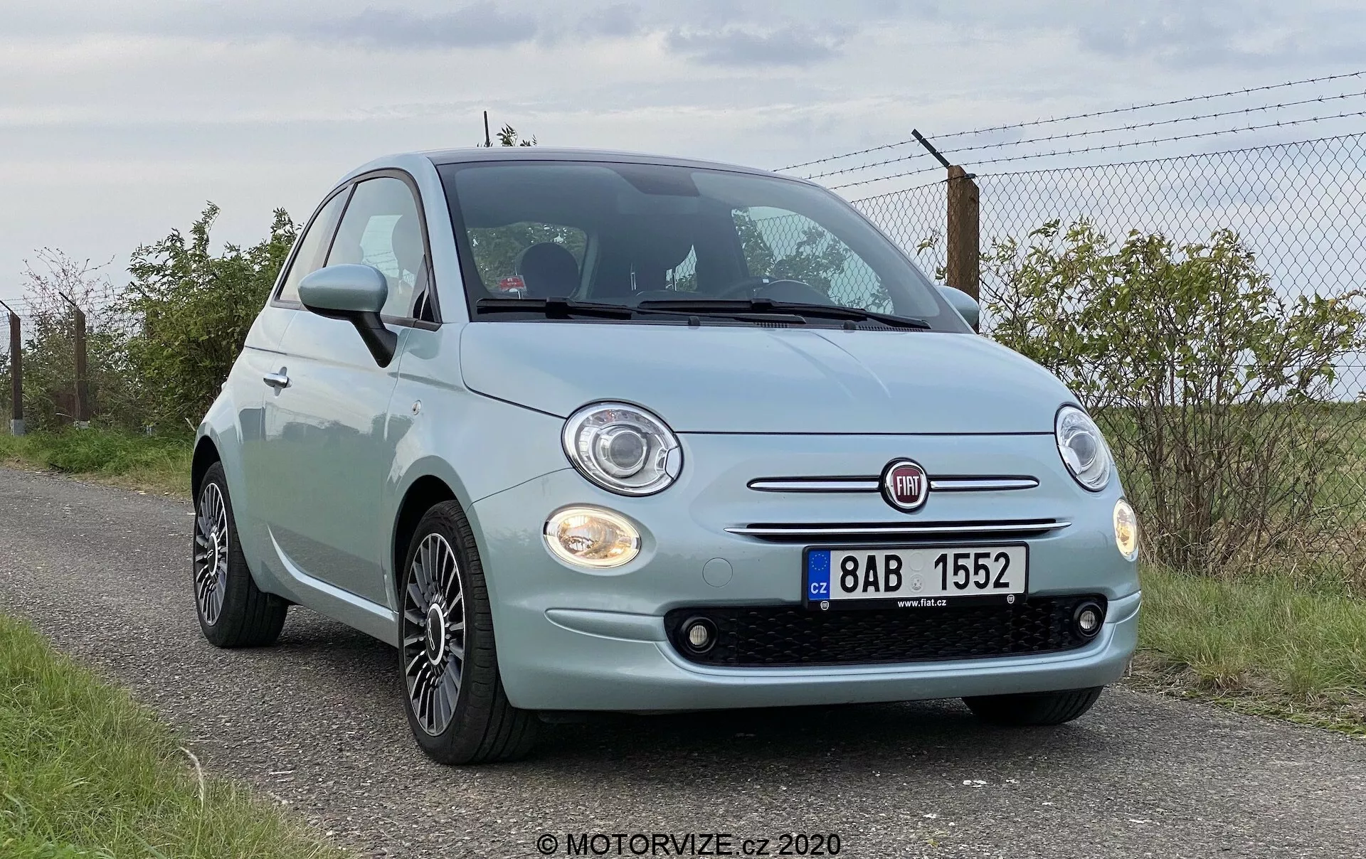 Nízký úhel předního pohledu na Fiat 500 z roku 2015, upozorňující na přední fasádu, kulatá světla, chromové detaily mřížky, mlhová světla a prominentní znak Fiat. Kapota auta má charakteristické křivky a přední nárazník obsahuje viditelné parkovací senzory. Viditelná jsou také litá kola. Vozidlo je zaparkováno na kraji cesty se zelenou plochou a plotem v pozadí, což naznačuje venkovské nebo polovenkovské prostředí.