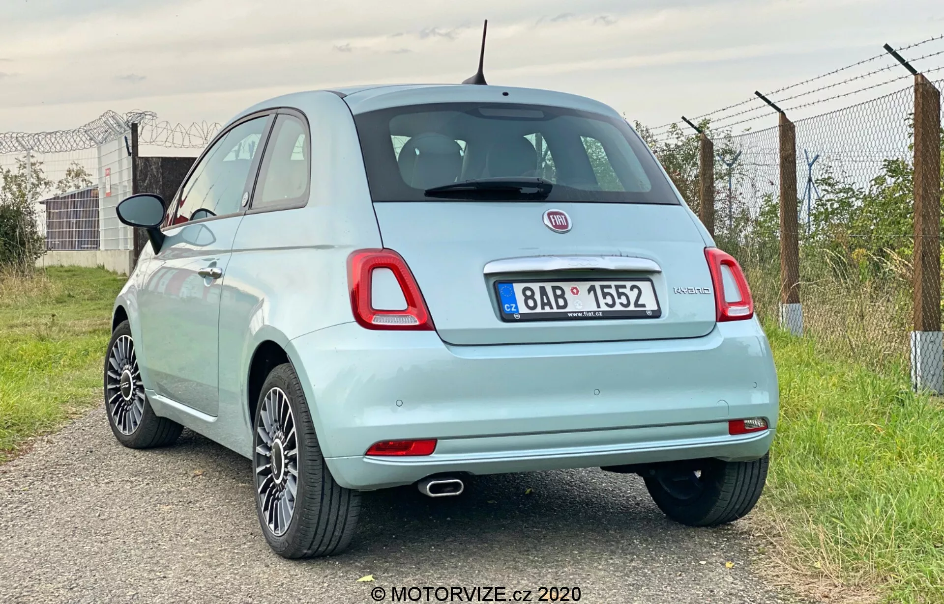 Obrázek zobrazuje zadní tříčtvrteční pohled na světle modrý hatchback Fiat 500 z roku 2015. Zaměřuje se na zadní a částečný profil na straně, pořízený z pravé zadní strany vozidla. Zvýrazňuje pravé zadní kolo, zadní světlo, částečný pohled na pravé zrcátko a dveře spolujezdce. Auto je umístěno mírně vlevo, s úhlem kamery mírně nižším než je linie střechy. Významné detaily zahrnují jedinou výfukovou trubku, litá kola, standardní žárovková zadní světla, odznaky značky a modelu, malý zadní spoiler, anténu ve stylu stožáru a viditelný uzávěr paliva. Prostředí působí venkovsky s otevřenou oblohou, zelení a plotem.