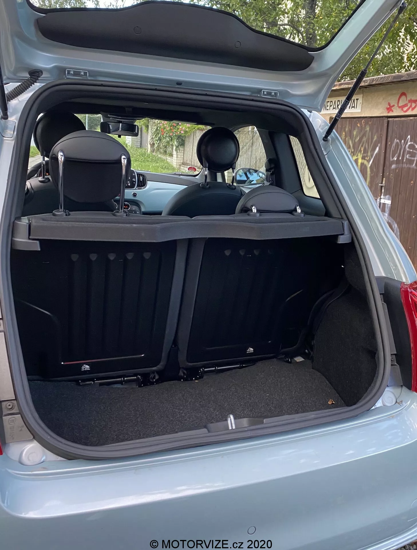 Obrázek ukazuje zadní interiér Fiat 500 2015 z vnějšího zadního úhlu s plně otevřenými dveřmi zavazadlového prostoru. Zavazadlový prostor je zobrazen z mírně mimo středového pravého úhlu, což odhaluje hloubku kufru a různé zadní interiérové prvky. Moderní čistý design zahrnuje kobercové povrchy, zatahovací žebrovaný kryt zavazadlového prostoru, dělená zadní sedadla s opěrkami hlavy a ochrannou lištu. Technologické prvky zahrnují vnitřní světlo, 12V napájecí zásuvku, plastové lišty, kruhové výřezy a boční panely.