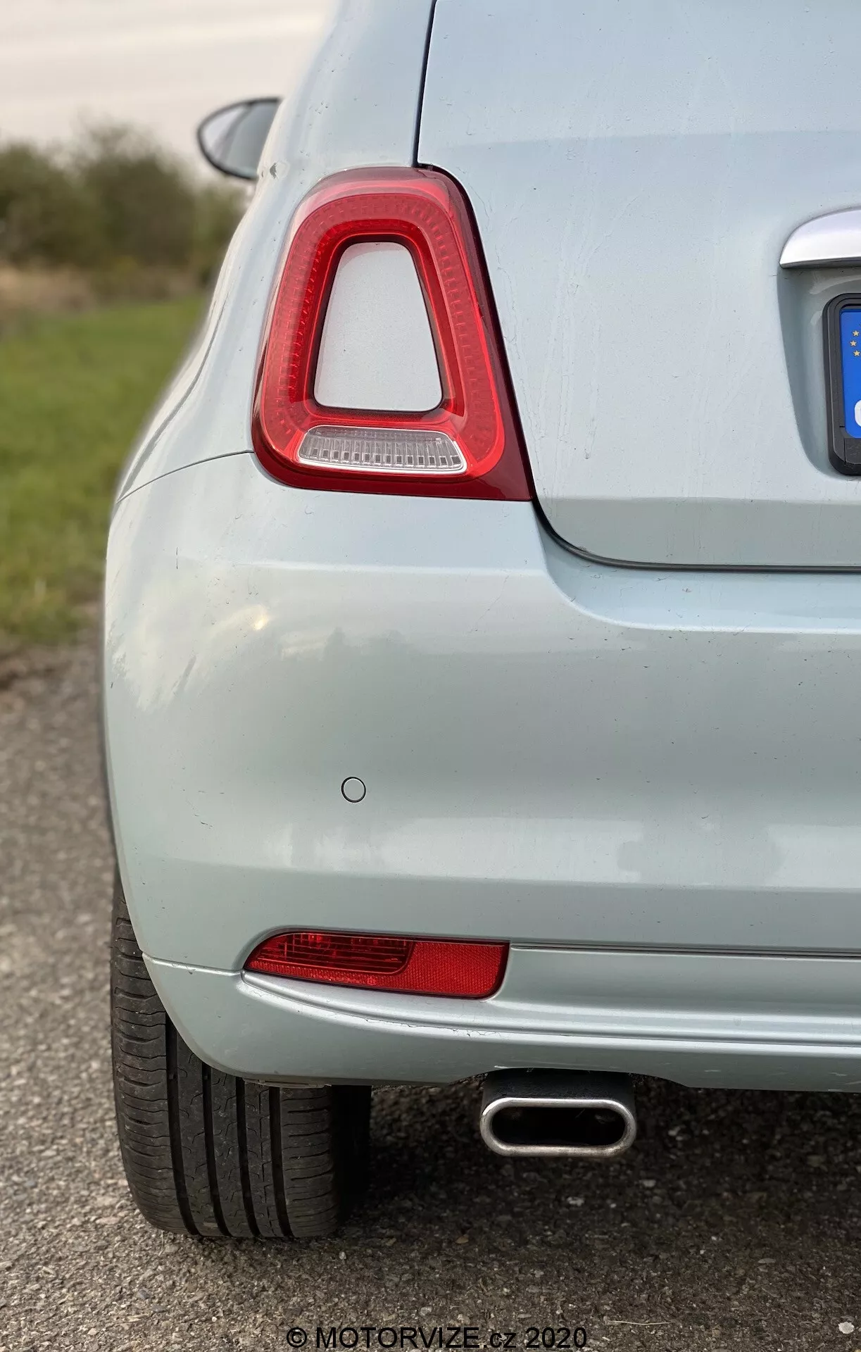 Blízký snímek zadní levé strany světle zbarveného vozu, zdůrazňující charakteristické zadní světlo s červeným krytem a technologií LED, červený reflektor, zapuštěný parkovací senzor a výfukovou trubku s chromovanou úpravou. Světle metalická barva a nízká hloubka ostrosti zdůrazňují čistý a dobře udržovaný stav vozidla.
