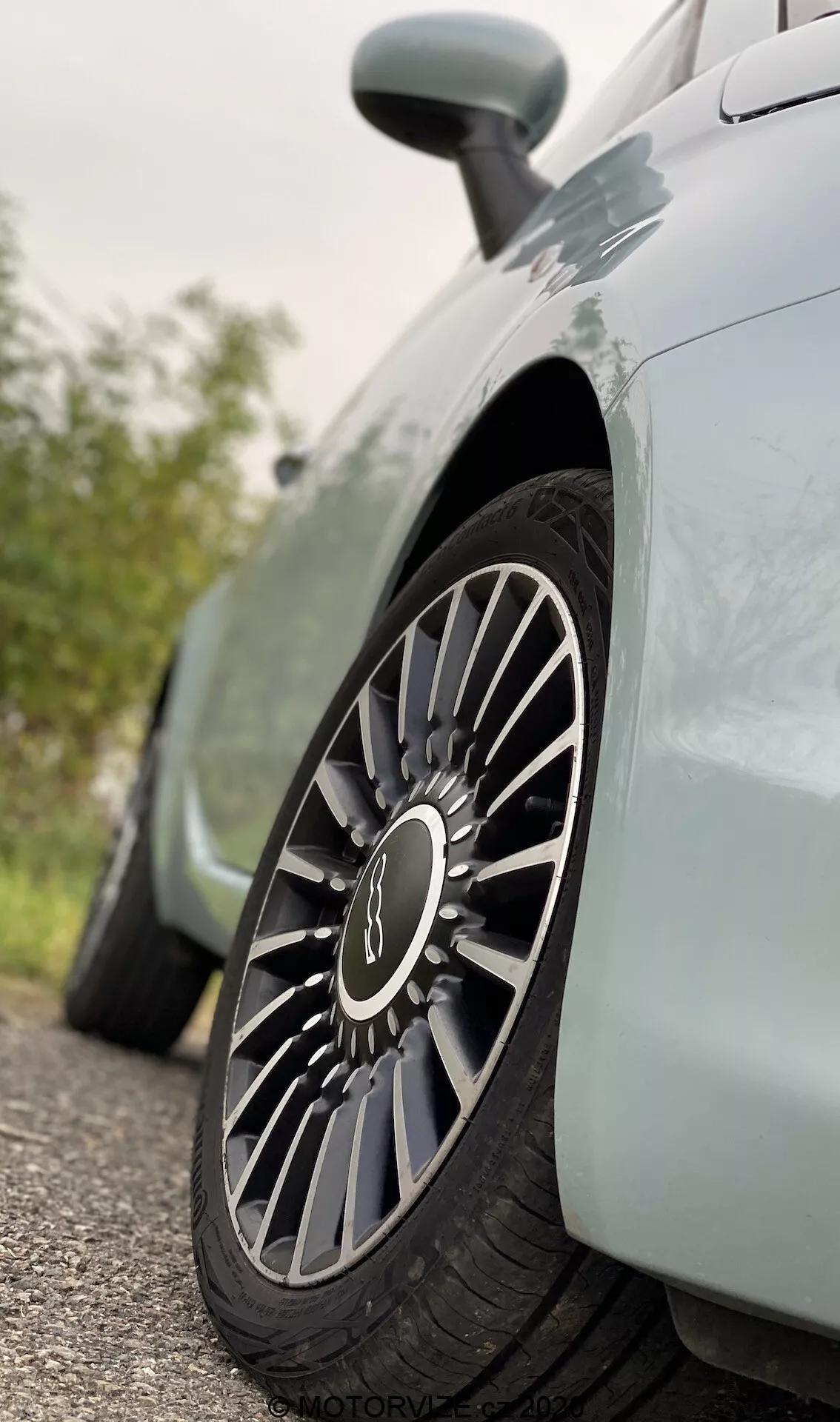 Detailní pohled na přední kolo a část předního blatníku Fiat 500 2015, snímaný z nízké perspektivy. Pozornost je zaměřena na vícepaprskové slitiny kola s černými a stříbrnými paprsky, které naznačují sportovní design. Středová krytka kola nese rozmazané logo, které naznačuje značku. Jsou viditelné detaily boční stěny pneumatiky, zatímco pozadí je měkce rozostřené, což zvýrazňuje dominanci kola a elegantní, aerodynamické linie těla vozu.