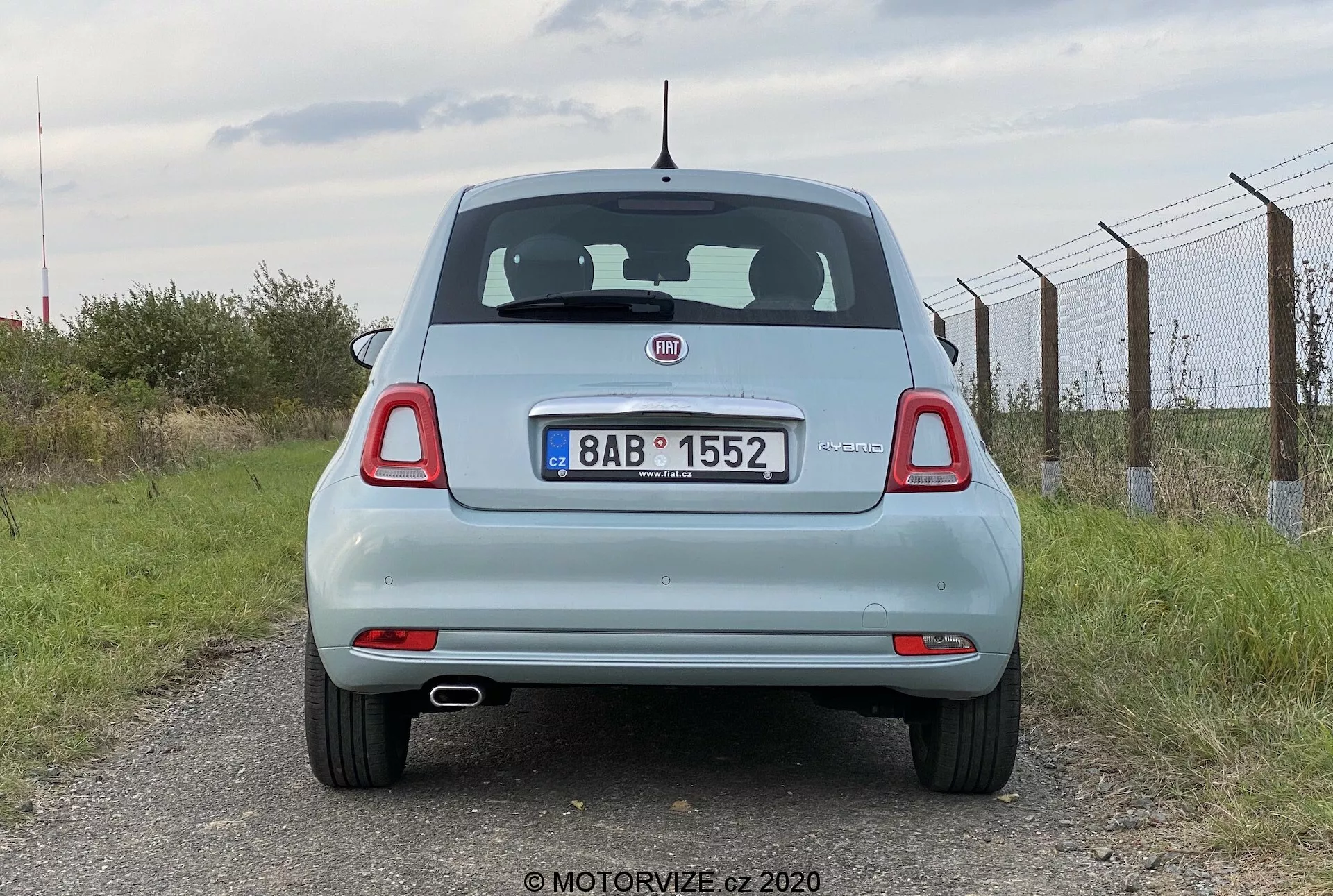 Zadní pohled na světle modrý automobil Fiat 500 2015. Perspektiva z úrovně očí zachycuje stojící vůz a ukazuje jeho celý zadek včetně kufru, zadního skla, zadních světel, zadního nárazníku a SPZ. Vlastnosti zahrnují výrazné logo výrobce, stěrač zadního skla, vysoko umístěné brzdové světlo, kulaté zadní světla s dalšími segmenty a SPZ uprostřed kufru pod logem nad rovnou zadním nárazníkem s integrovanými odrazkami. Krátká anténa je umístěna uprostřed střechy směrem dozadu. Zamračená obloha a venkovní okolí s plotem vpravo a trávou pod ním.