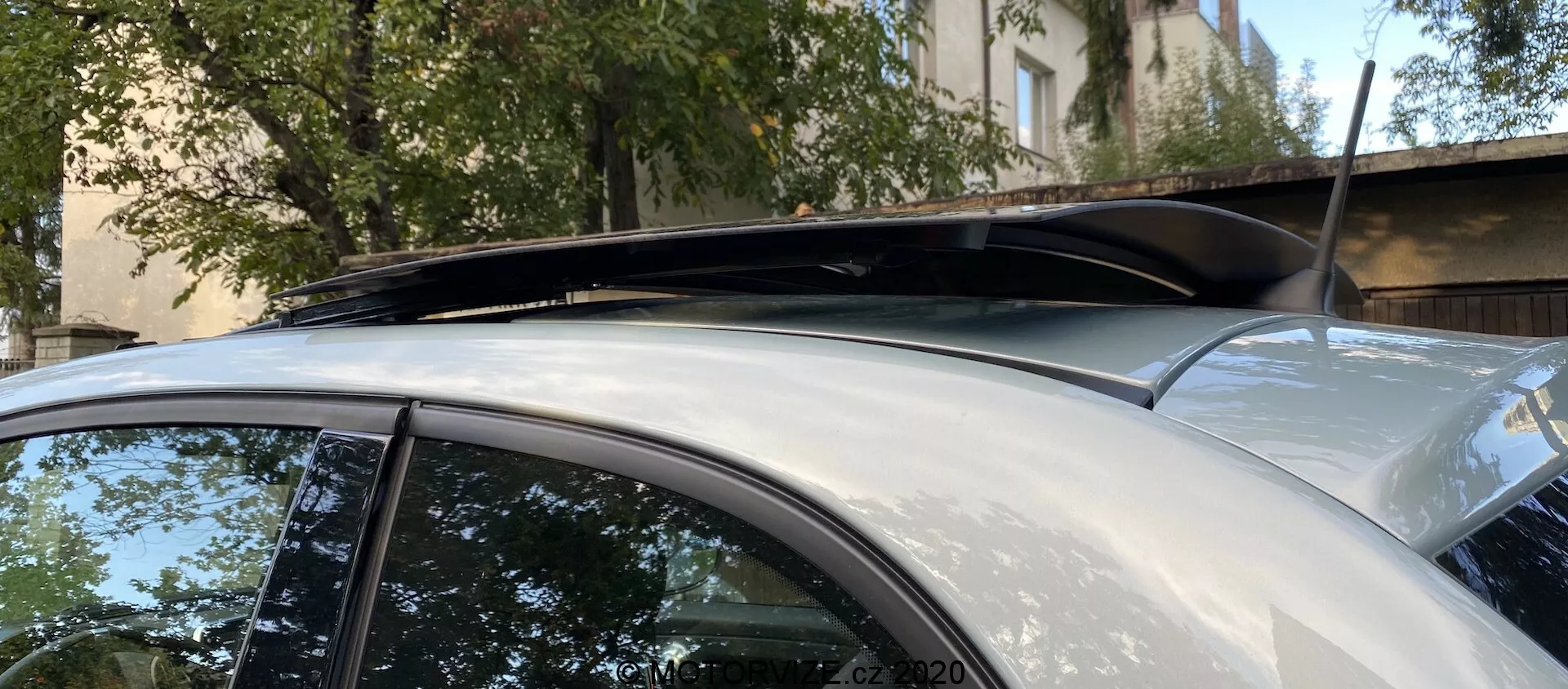 Detailní pohled na střešní část Fiat 500 z roku 2015, vybavený panoramatickým střešním oknem s tónovaným sklem a střešními nosiči. Obrázek zdůrazňuje vysokou úroveň výbavy a praktické detaily, zachycený z boční perspektivy se zaměřením na horní část vozidla. Stříbřitošedý lak vozu je v dobrém stavu bez viditelných škrábanců nebo promáčklin.