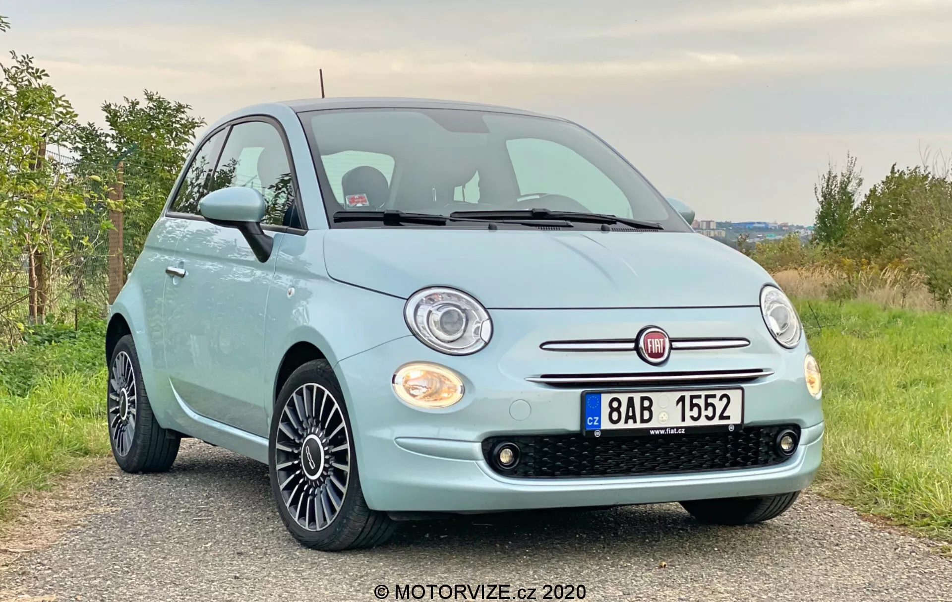 Přední tříčtvrteční pohled na světle modrý Fiat 500 z roku 2015 s chromovanou mřížkou a kulatými halogenovými světlomety v mírně nízkém úhlu. Obrázek zvýrazňuje přední část, mřížku, světlomety, kapotu a slitá kola s černým vícepaprskovým designem. Auto je zaparkováno na štěrkové nebo prašné cestě v venkovské oblasti se zelenou vegetací v pozadí.