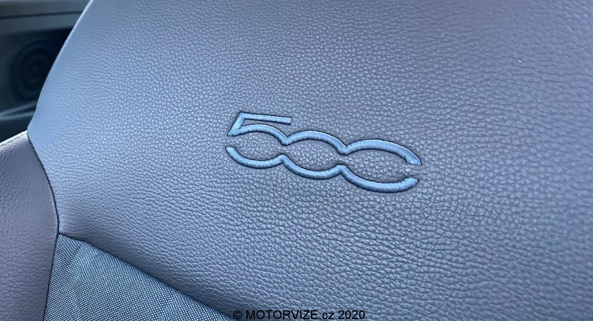 Detailní záběr zobrazující stylizované logo '500' na přístrojové desce interiéru Fiat 500 2015. Kožená textura a přesné prošívání panelu jsou zvýrazněny osvětlením a úzkou hloubkou ostrosti, čímž se zdůrazňuje vysoká kvalita řemeslného zpracování interiéru vozidla.