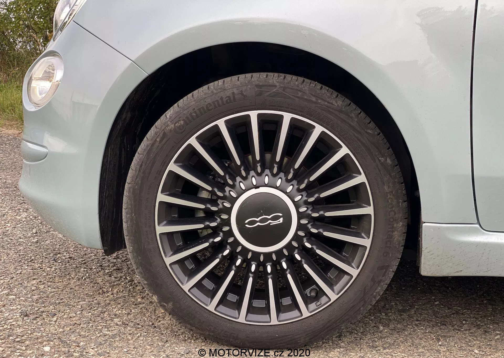 Blízký pohled na přední kolo z řidičovy strany vozu Fiat 500 2015, který zobrazuje litý ráfek s tenkými, vícebarevnými paprsky sbíhajícími se k emblému ve středové čepici. Pneumatika, s jasně viditelným dezénem a textem na bočnici, je bezpečně připevněna, přičemž nad kolem je vidět světle zbarvená karoserie a křivka blatníku.