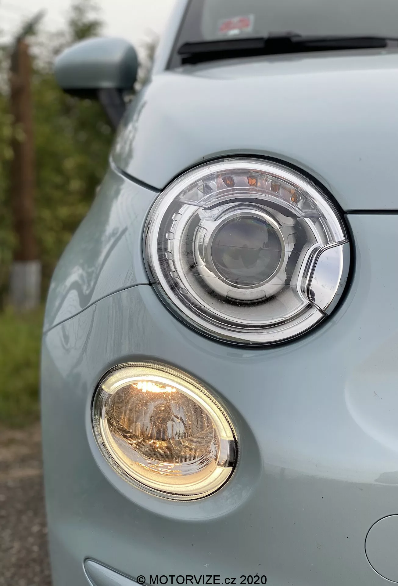 Detailní pohled na levý přední roh světle modrého Fiatu 500 2015, zaměřující se na halogenový projektorový světlomet v kruhovém designu obklopeném chromovaným kroužkem. Pod ním menší mlhové světlo vyzařuje teplé, žlutavé světlo. Obrázek z nízkého úhlu zvýrazňuje hladký a zaoblený přední nárazník s výraznou mřížkou a oblastí pro registrační značku, podtrhující dobře udržovaný, městský design vozu.