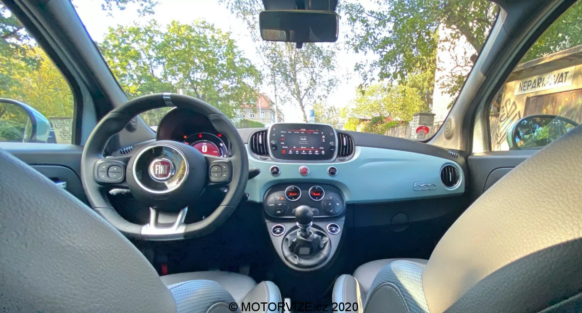 Fiat 500 2015 interiér, pohled ze zadního sedadla, ukazující přední palubní desku, volant s emblémem Fiat, částečně viditelný přístrojový panel, základní infotainment systém s tlačítky, otočné ovladače klimatizace, dvoubarevná palubní deska, světle šedé látkové sedačky, dveře pro spolujezdce s klikou a mřížkou reproduktoru, jednoduchý a funkční design bez pokročilých technologických prvků.