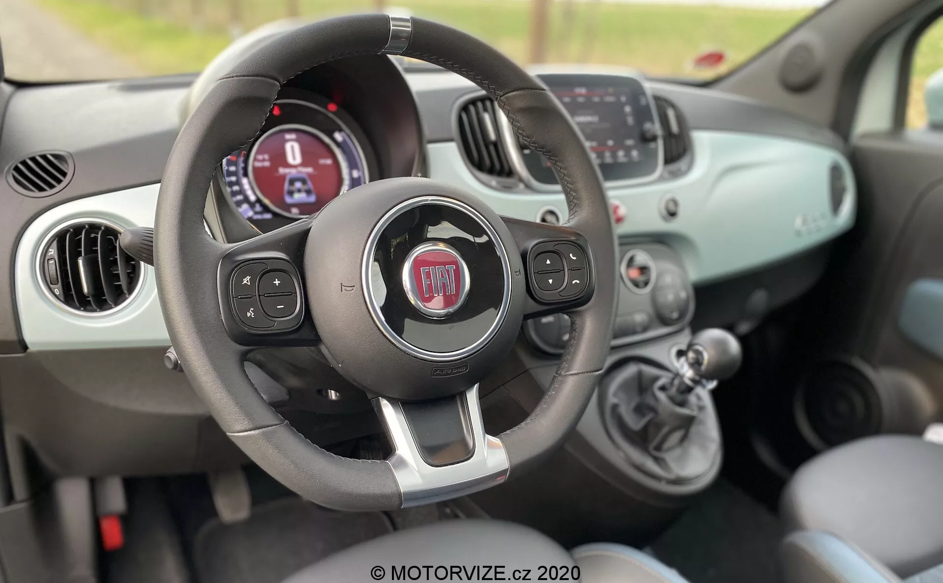 Interiér vozu Fiat 500 z roku 2015 zachycený z pohledu spolujezdce, ukazující volant s multifunkčními tlačítky, přístrojový panel s digitálním displejem, manuální klimatizaci a řadící páku.