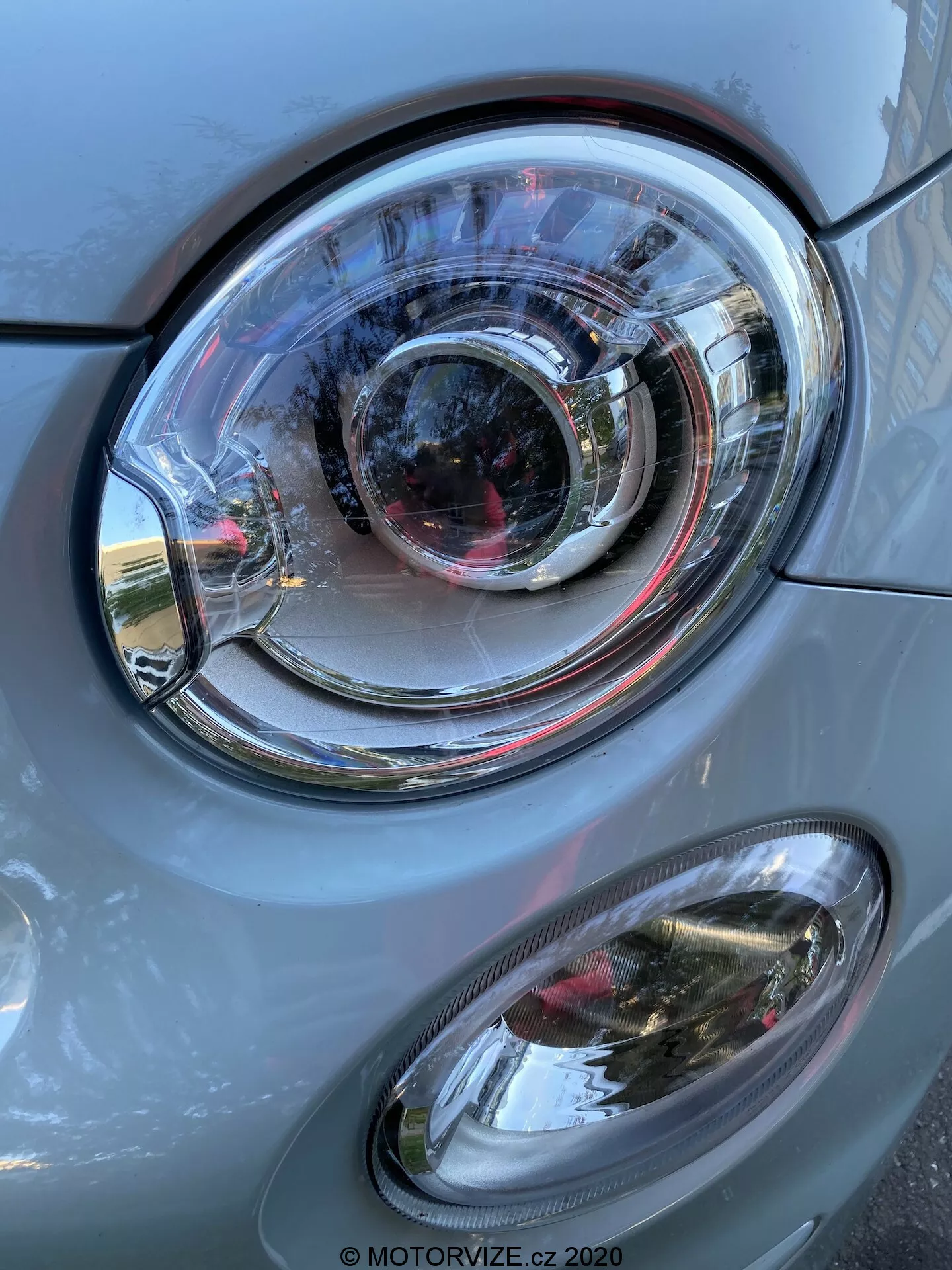 Detailní pohled na přední levý bok vozu Fiat 500 2015 se zaměřením na přední světlomet. Obrázek, ve vertikální orientaci, zobrazuje výrazný plastový světlomet s průhlednou čočkou a složitým designem soustředných kruhů, což naznačuje projektorový typ světlometu a možná i adaptivní osvětlovací technologii. Pod hlavním světlometem se nachází oválná mlhovka s průhledným krytem. Karoserie vozu je šedá s kovovým leskem, dokonale zpracovaná a odrážející denní světlo a prvky okolního prostředí. Přední světlomet a mlhovka jsou ve výborném stavu, což naznačuje dobře udržovaný nebo nový vůz.