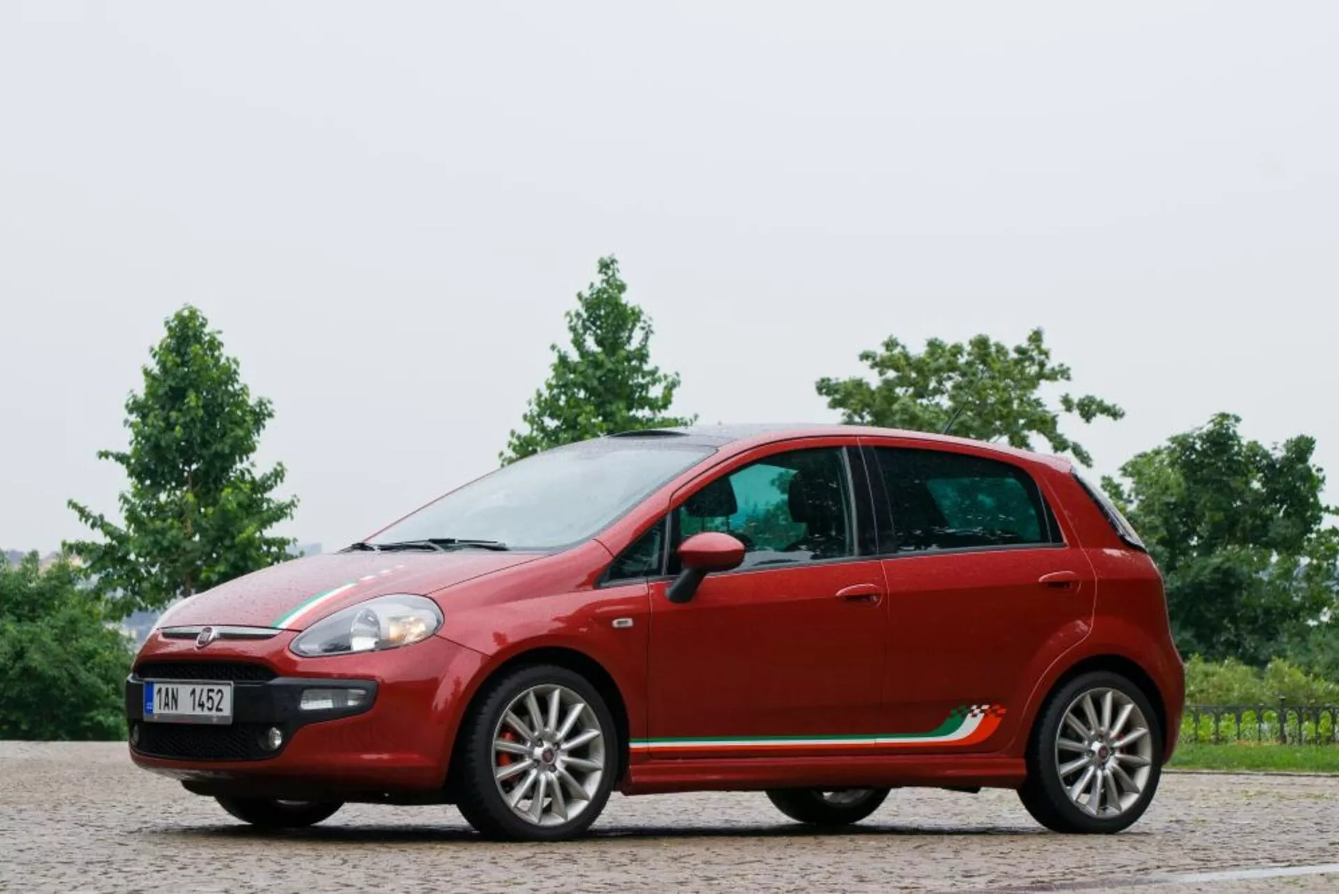 Plný boční profil červeného hatchbacku Fiat Punto z roku 2011 s zakřivenou střechou, konfigurací pěti dveří s litými koly, bočními zrcátky na dveřích, výraznými liniami a plastovým lemováním na štěrkovém povrchu s pozadím zeleně.