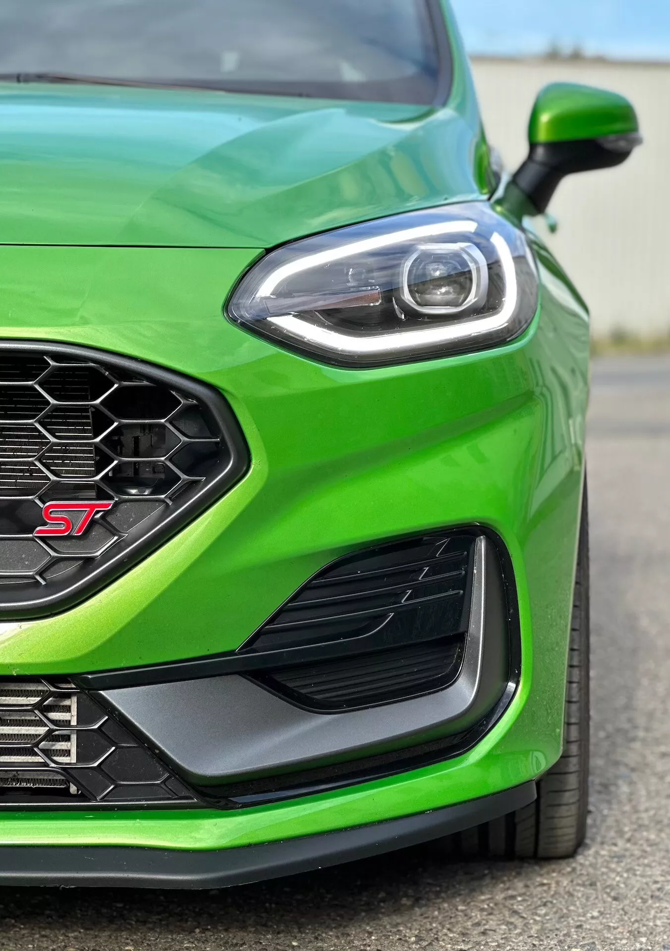 Detailní pohled na zelený Ford Fiesta ST 2022 zaměřený na výkonnostní přední levý kvadrant, zdůrazňující sportovně laděný trim, mřížku s včelím pláštěm pro optimální proudění vzduchu, pokročilý design LED světlometů, aerodynamické boční otvory a přední nárazník pro lepší kanalizaci vzduchu.