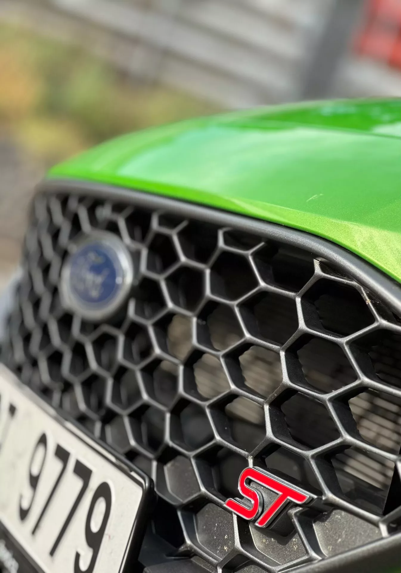 Detailní pohled na část přední části Fordu Fiesta ST 2022 s hexagonální mřížkou, odznakem ST a logem značky v ostrém zaostření s zelenou kapotou, zdůrazňující vlastnosti pro výkon a design