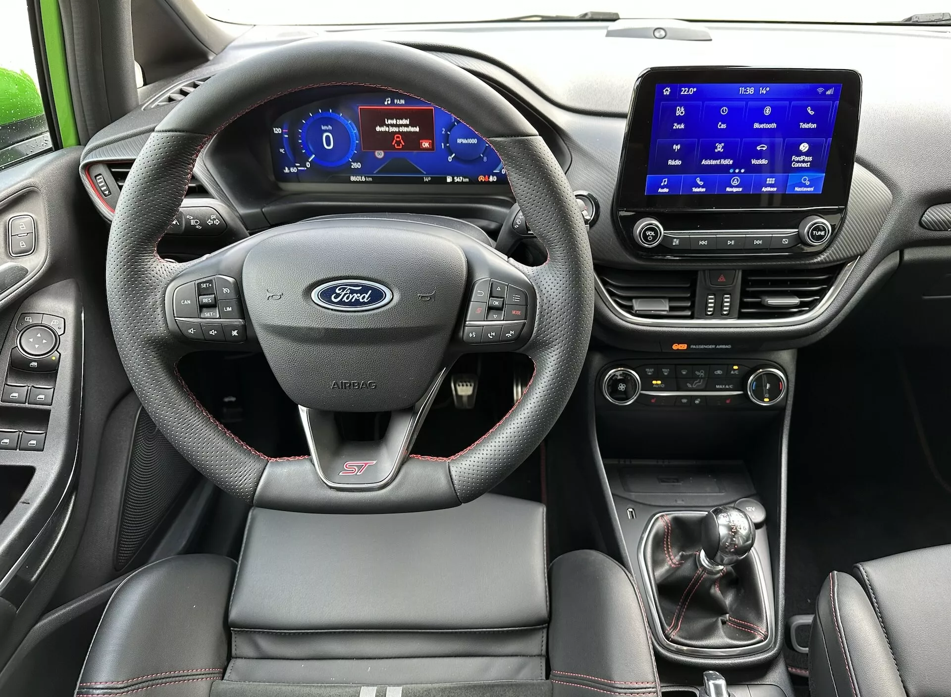 Interiér Ford Fiesta 2022 z pohledu zadního sedadla, zdůrazňující volant s logem Ford a odznakem ST, digitální přístrojový panel, dotykový systém infotainment, manuální řadící páku a sportovní sedadlo řidiče s kontrastním prošíváním.