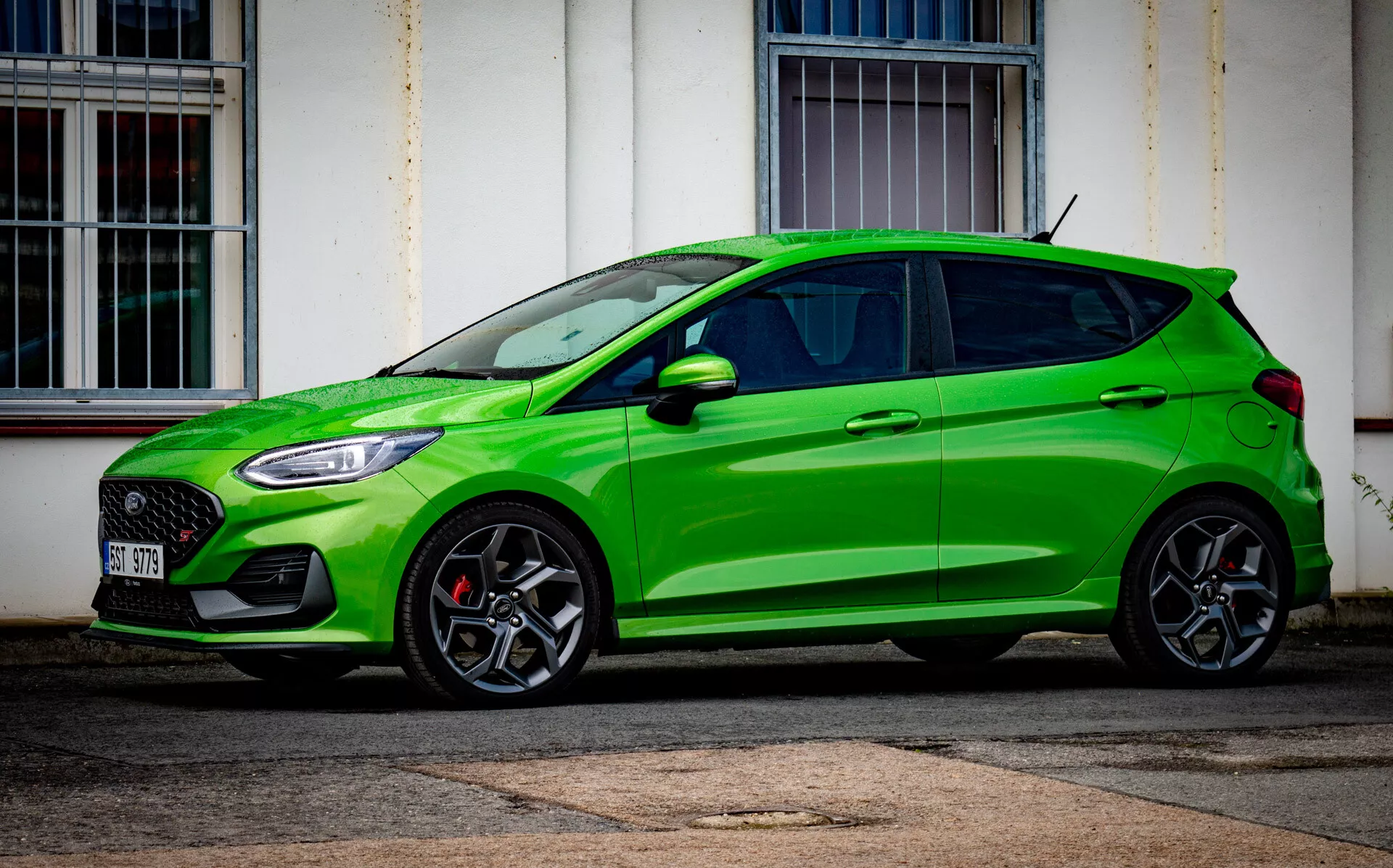 Ford Fiesta 2022 ve světle zelené barvě, pohled z mírného předního úhlu ukazuje sportovní boční profil, design pětidveřového hatchbacku, dvoubarevná kola z lehké slitiny s více paprsky, výkonnostní prvky včetně červených třmenů brzd a včelí plástve na mřížce, ostré LED světlomety, aerodynamické prahy a tónovaná okna, zaparkovaný v městském prostředí