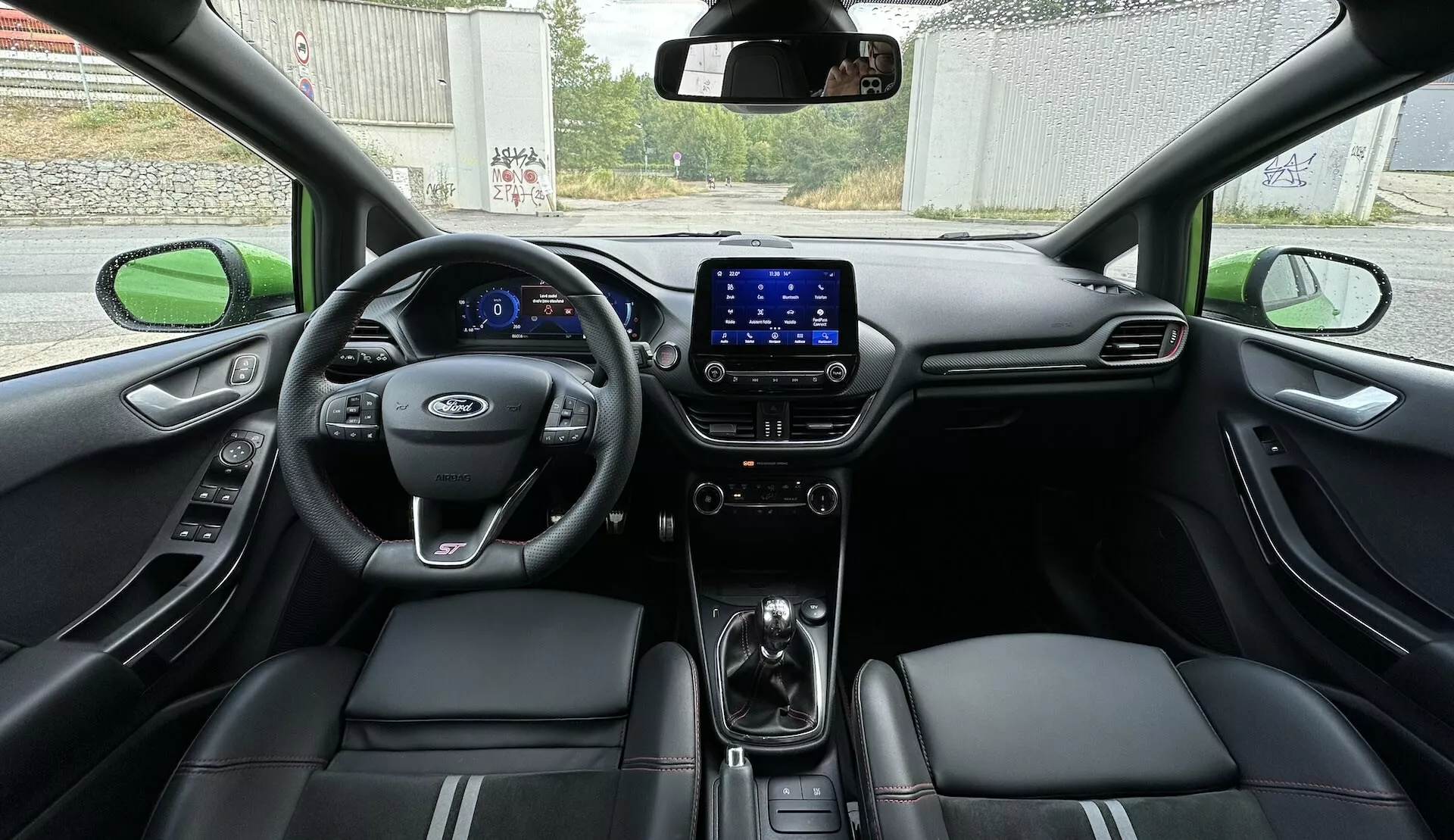 Interiér Ford Fiesta 2022 z pohledu ze zadního sedadla, ukazuje celý kokpit s digitálním palubním stolem, multifunkčním volantem a koženými sedadly s vyšíváním