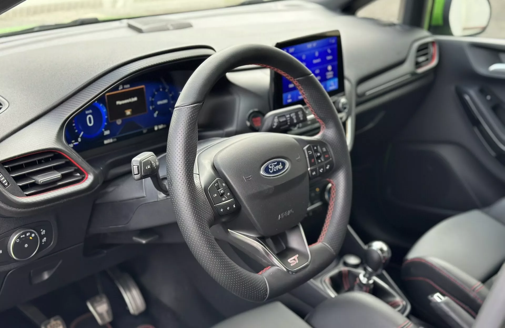 Interiér Ford Fiesta 2022 z pohledu řidiče s koženým volantem s ovládacími tlačítky, logem Ford, digitálním přístrojovým panelem a moderním informačním systémem na palubní desce