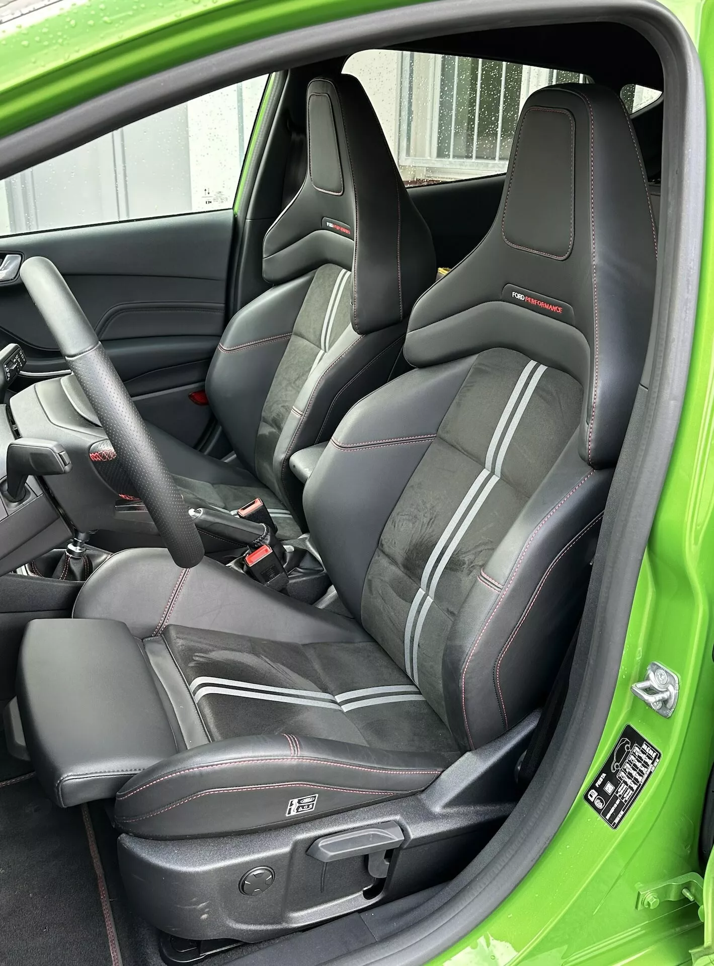 Detailní pohled na sportovní kožené sedadlo řidiče s červeným lemováním, trojbodový bezpečnostní pás a elektrické ovládání sedadla, doplněné o zelenou výzdobu interiéru u Ford Fiesta 2022.