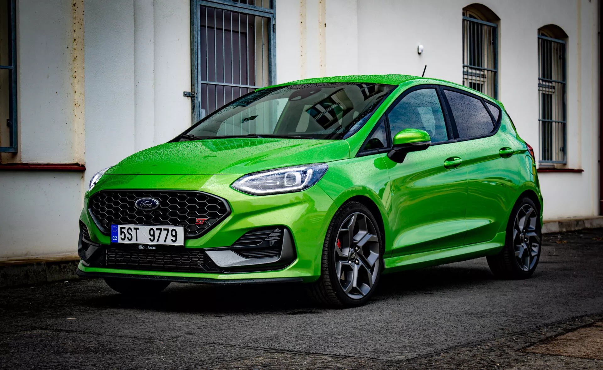 Ford Fiesta 2022 v jasné zelené barvě, předvádějící svůj sportovní design z nízkého úhlu pohledu na přední část zdůrazňující přední nárazník, mřížku, světlomety, kapotu, blatník, kola a odraz čelního skla na městské ulici po dešti