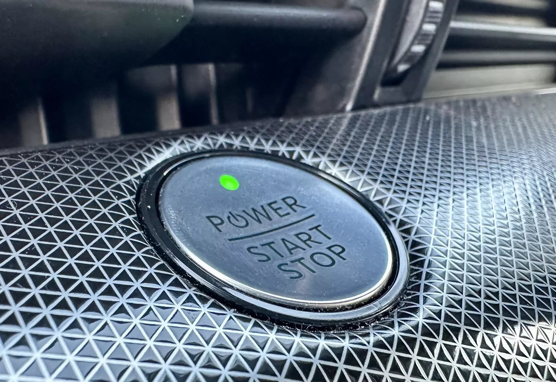 Detailní pohled na tlačítko pro spuštění/zastavení motoru ve Ford Mustang-Mach-E 2020, s textem 'POWER START STOP', zelenou kontrolní lampičkou a texturovaným okolním designem