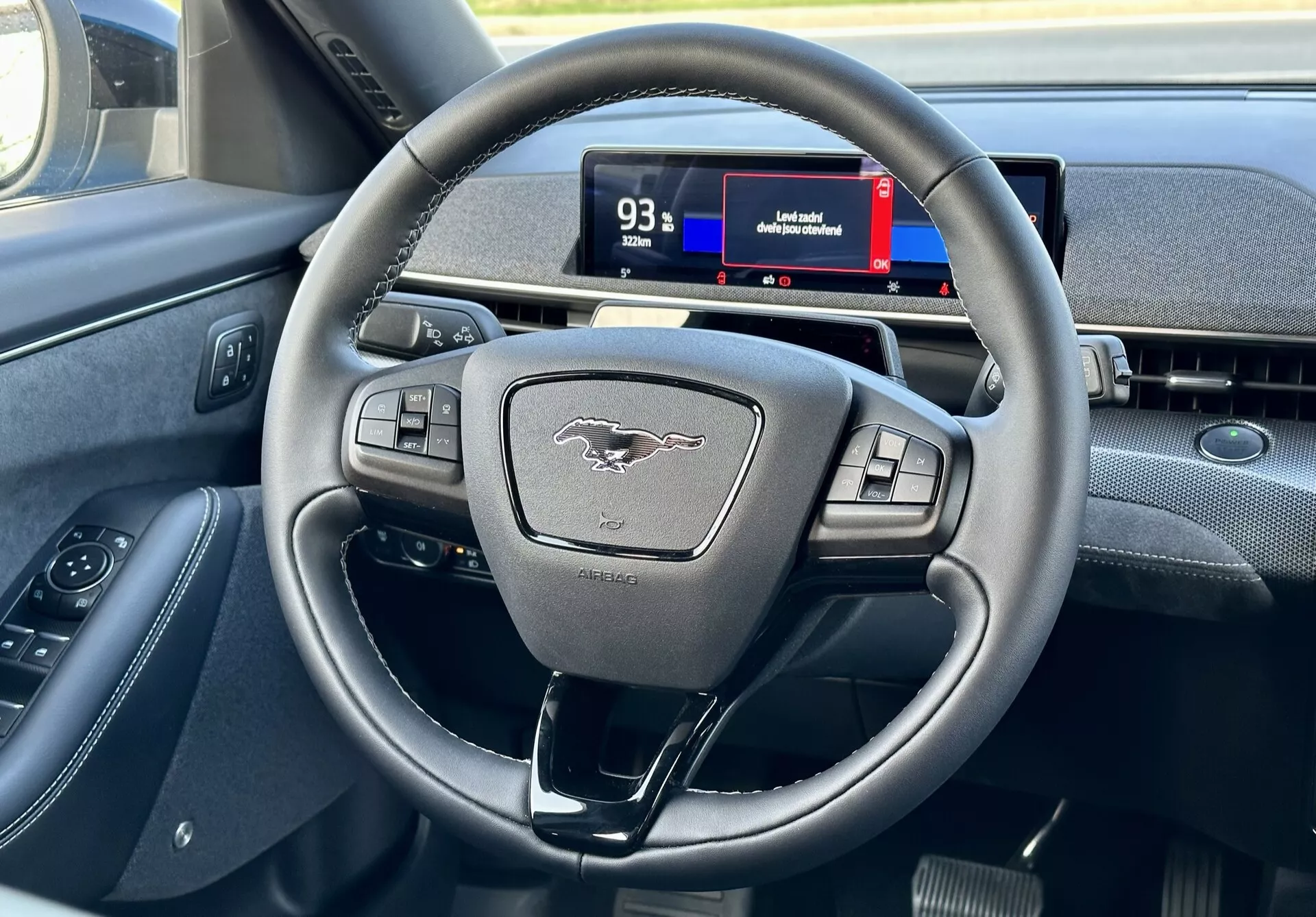 Interiér Ford Mustang Mach-E 2020 zdůrazňující moderní tříramenný volant s multifunkčními tlačítky a digitální přístrojovou deskou z pohledu řidiče, ukazuje ovládací prvky, emblém a náznaky navigace.