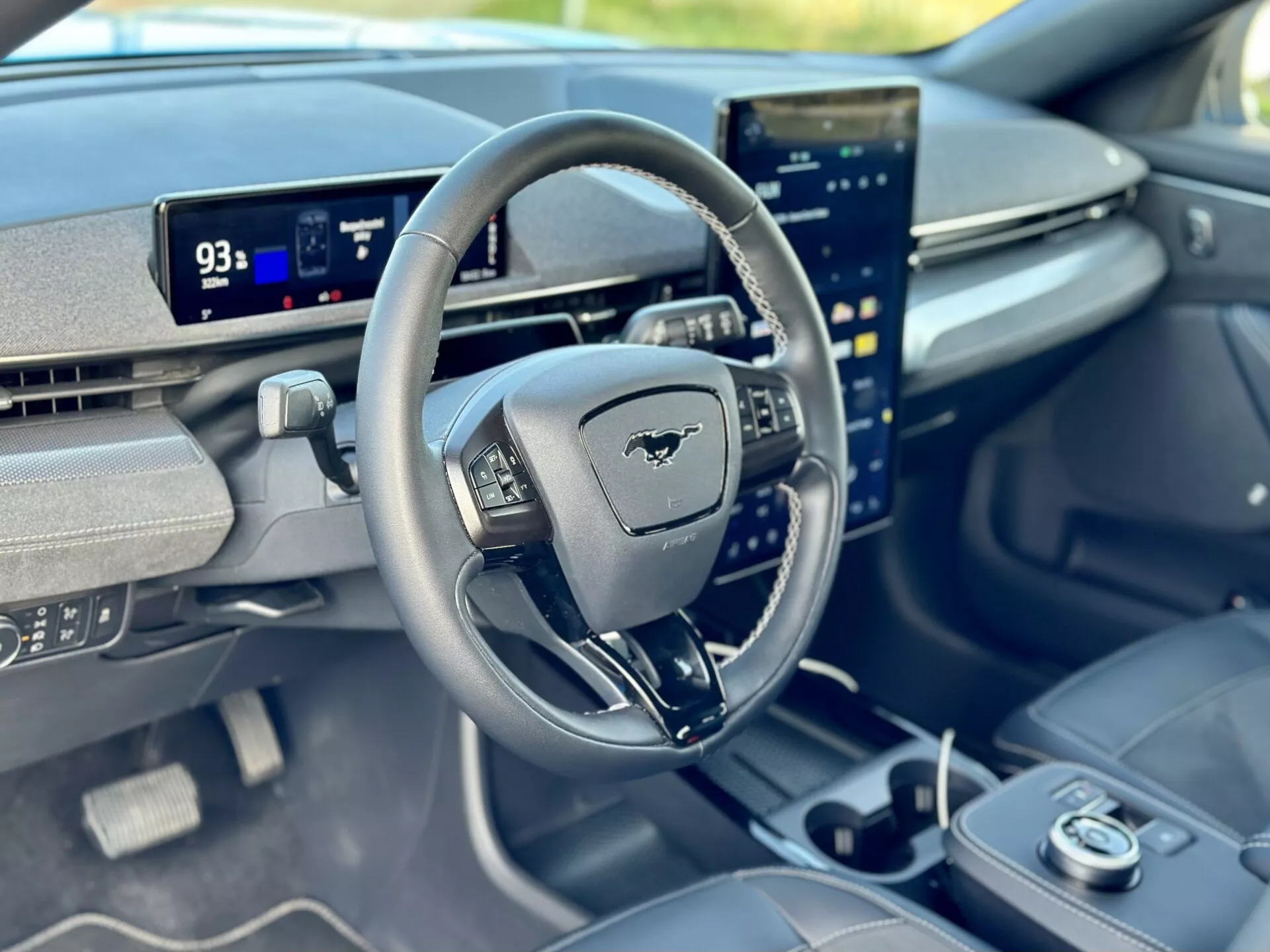 Vnitřní pohled na Ford Mustang Mach-E 2020 zaměřený na kokpit řidiče z pohledu předního spolujezdce, s volantem s emblémem Mustang, digitálním palubním počítačem v režimu Park s dojezdem 93 mil a svisle orientovaným centrálním dotykovým displejem integrovaným s pokročilými možnostmi informačního a zábavního systému.