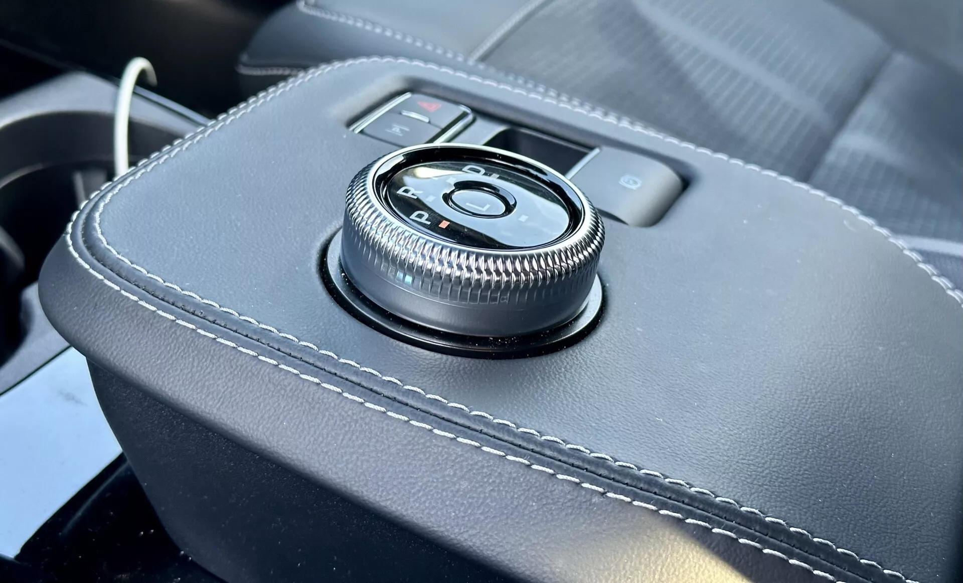 Detailní pohled na interiér Ford Mustang Mach-E 2020 s otočným ovladačem na centrální konzoli na povrchu obaleném kůží s bílým prošíváním, poskytující ergonomické rozhraní pro informačně-zábavní systém