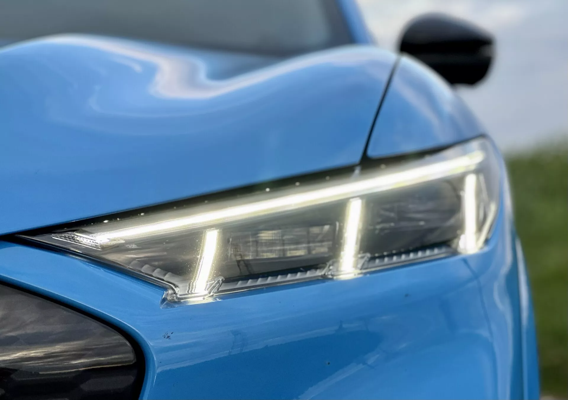 Detailní pohled na přední levou část Ford Mustang-Mach-E 2020 s důrazem na elegantní LED světlomet, živou modrou barvu, dynamické linie karoserie a část předního nárazníku a boční zrcátko, což zdůrazňuje moderní design a estetiku.