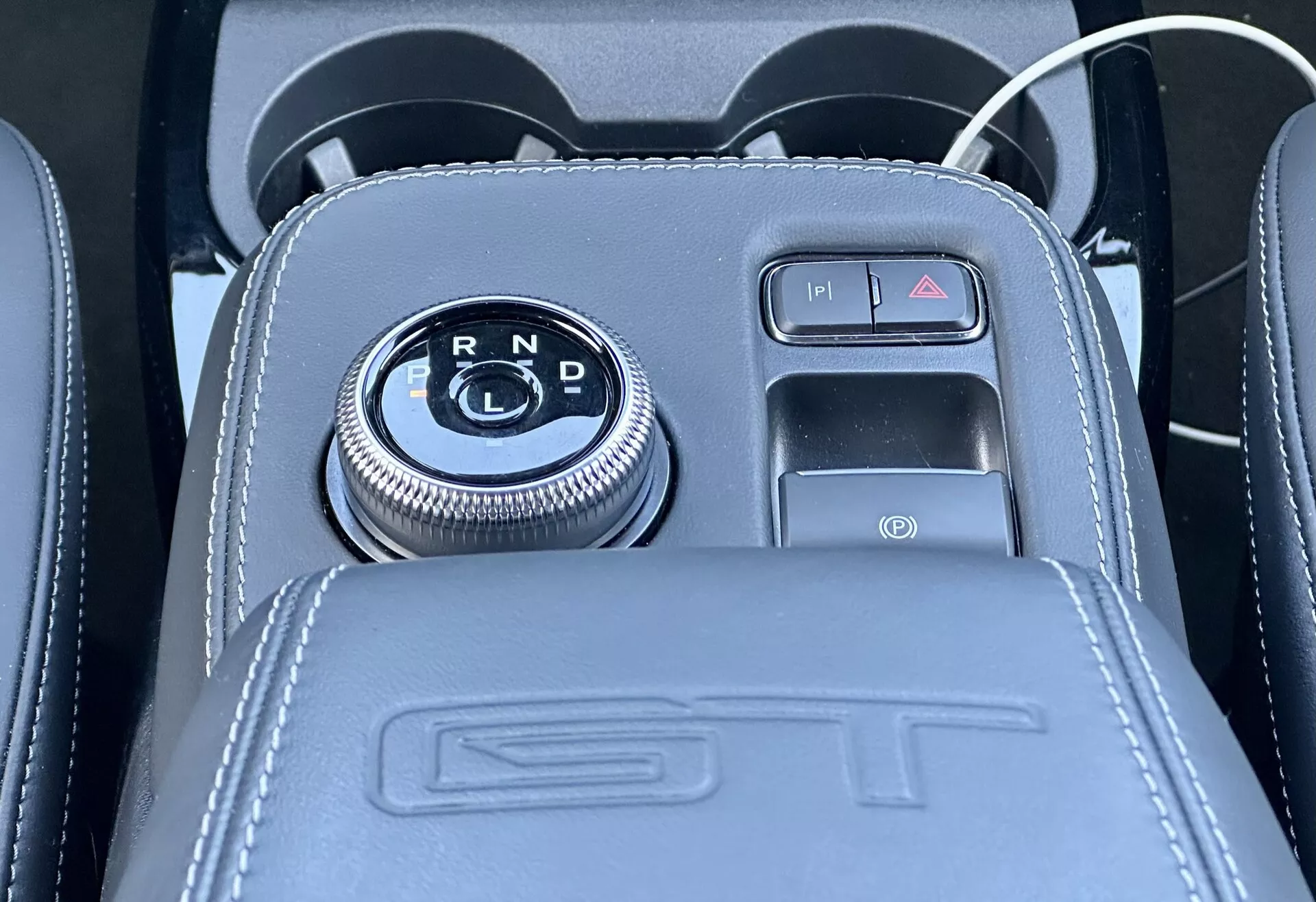Detailní pohled na středovou konzoli Ford Mustang Mach-E 2020 s otočným voličem převodovky umístěným v lesklém černém panelu s ukazateli zařazení R, N, D, obklopeným chromovým obložením, vedle tlačítkové elektronické parkovací brzdy s detaily potahů z kůže, reliéfním nápisem ST, a včetně portu USB a 12V zásuvky pro příslušenství