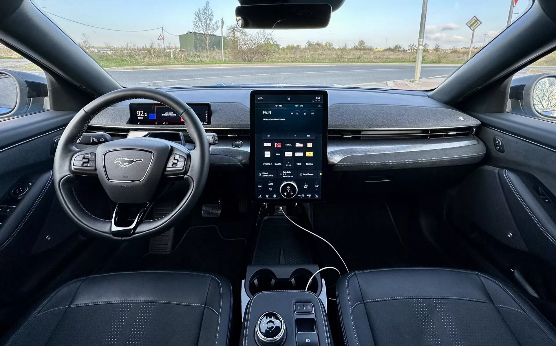 Pohled z první osoby na interiér Ford Mustang Mach-E 2020, zobrazující volant na levé straně s plochým dnem a emblémem, digitální přístrojový panel, velký vertikální dotykový informační systém na středové konzole s ovládacími knoflíky klimatizace níže, USB porty a luxusní čalounění v tmavých tónech s detailním prošíváním, naznačující pokročilé bezpečnostní a asistenční funkce, s viditelným venkovským pozadím skrze čelní sklo.