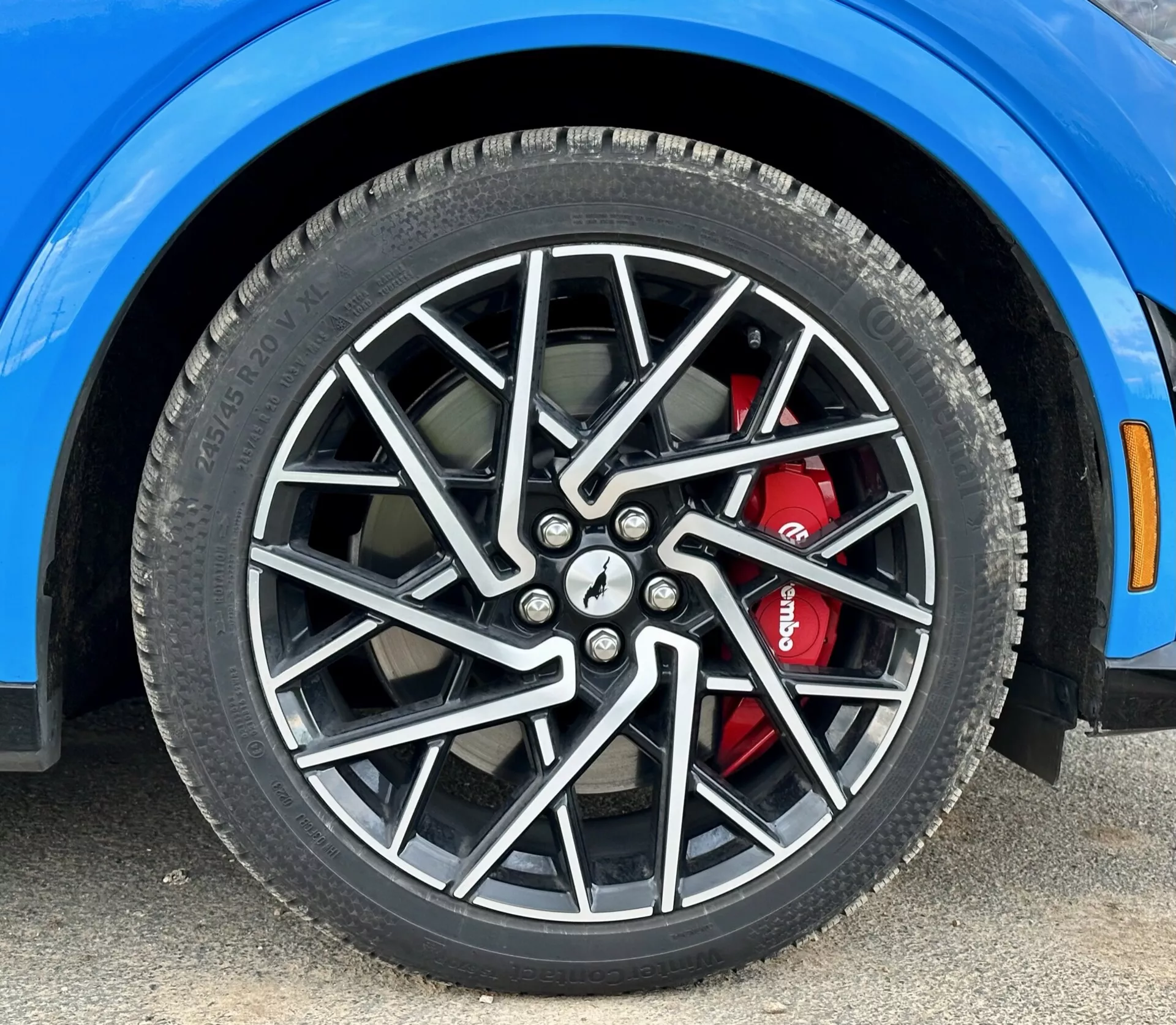 Detailní pohled na přední pravé kolo Fordu Mustang Mach-E z roku 2020 s vícepaprskovým slitinovým ráfkem a vysokovýkonným brzdovým systémem s červeným třmenem, zobrazující detaily celoroční pneumatiky a segment odvážně modrého blatníku, snímek z nízkého úhlu.