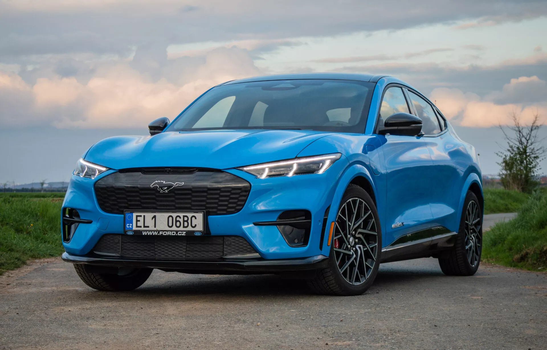 Modrý Ford Mustang Mach-E 2020 fotografován z nízkého a mírně excentrického úhlu, zobrazující mřížku, světlomety, kapotu a profil vozu včetně předního kola a blatníku, což zdůrazňuje jeho výkon a aerodynamický design na svítání nebo soumraku.