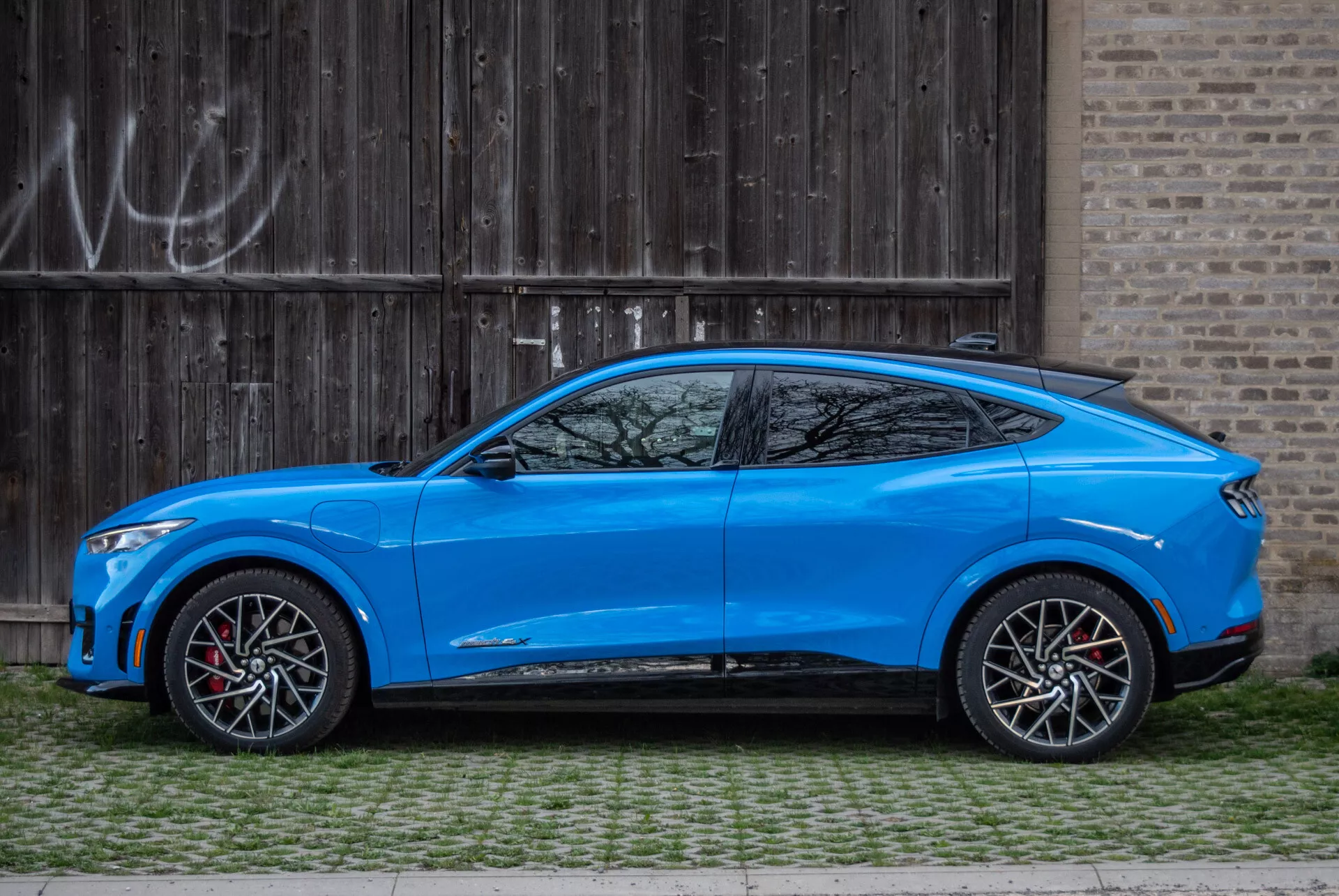 Modré crossover SUV Ford Mustang Mach-E 2020 zaparkované rovnoběžně s rustikálními dřevěnými garážovými dveřmi s graffiti, boční pohled ukazující aerodynamický design, skloněnou střechu ve stylu kupé, čtyřdveřový model s integrovanými klikami, aerodynamicky stylizované boční zrcátka s integrovanými směrovými signály, velká dvoubarevná slitinová kola s červenými brzdovými třmeny, střední světlou výšku, konturované karoserie se sochařskými liniemi a výraznými koly, zúženou linii oken s malým čtvrtým oknem za sloupkem C a sportovní prahy