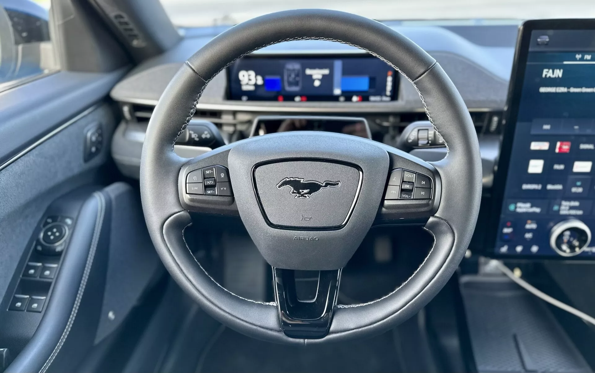 Pohled z interiéru Ford Mustang-Mach-E 2020 z perspektivy řidiče, obsahující tříramenný volant s ovládáním, digitální přístrojovou desku a velký dotykový informačně-zábavní displej