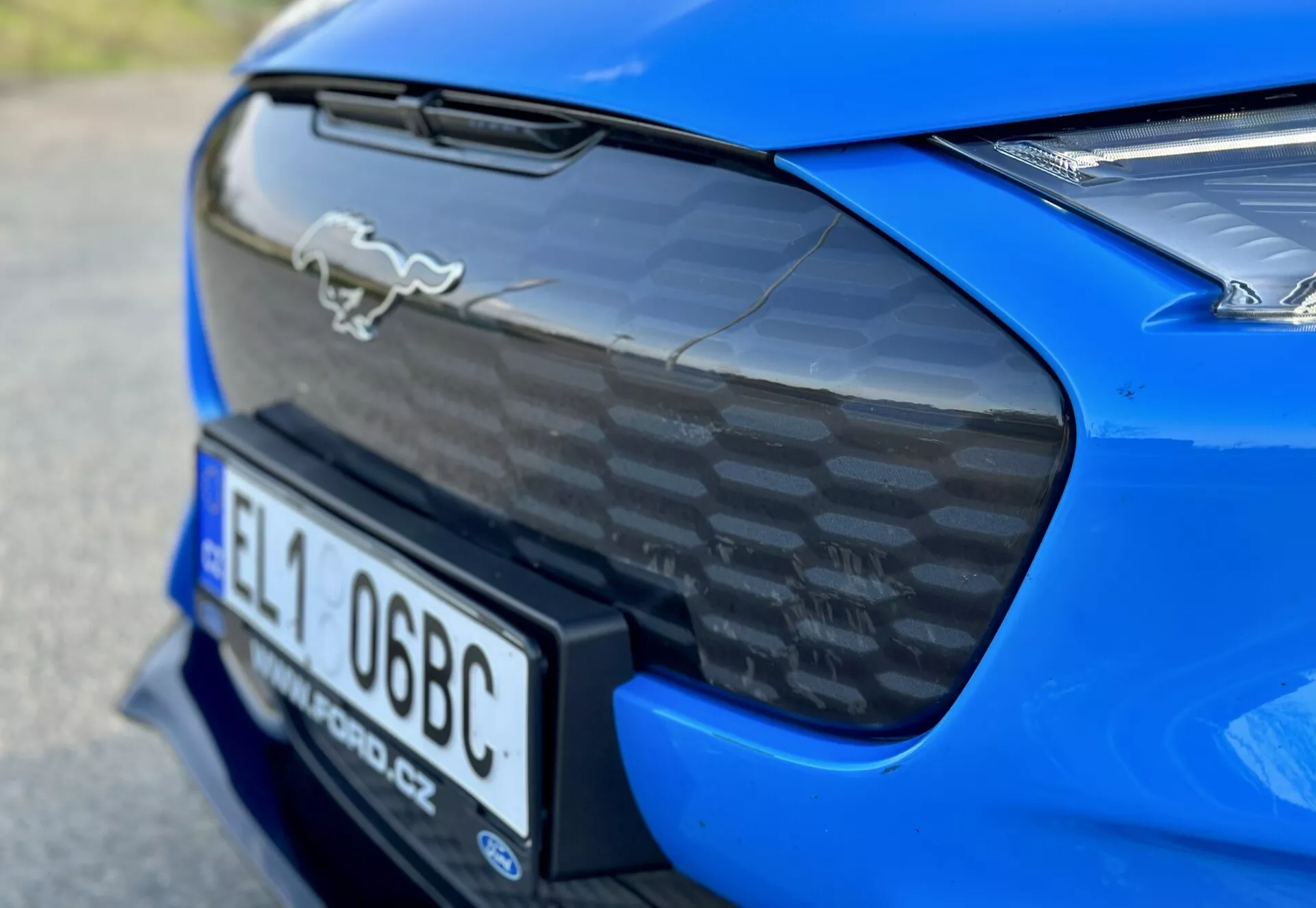 Detailní záběr na zadní část modrého sportovního auta Ford Mustang Mach-E 2020 ukazující chromový emblém koně, spoiler z uhlíkových vláken, integrovanou niky pro registrační značku a částečný pohled na LED zadní světla, zdůrazňující aerodynamické a výkonnostní vlastnosti auta.