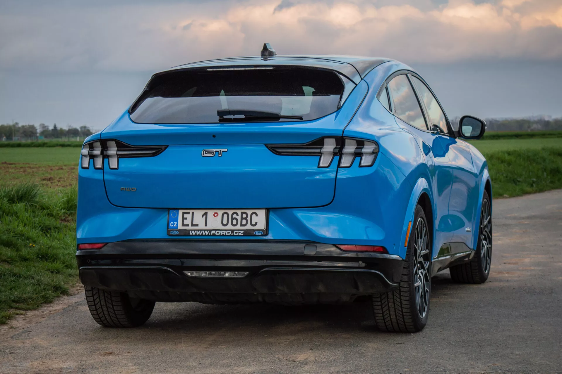 Ford Mustang-Mach-E 2020 fotografovaný zezadu a ze tříčtvrtečního úhlu, zdůrazňující jeho charakteristický design, včetně horizontálních LED zadních světel, emblému výrobce, integrovaného zadního spoileru, zadního nárazníku vybaveného senzory bez viditelného výfuku naznačujícího status elektromobilu, evropské registrační značky, slitinových kol s více paprsky, živé modré barvy a aerodynamického tvarovaného karosérie, na pozadí venkovské krajiny.